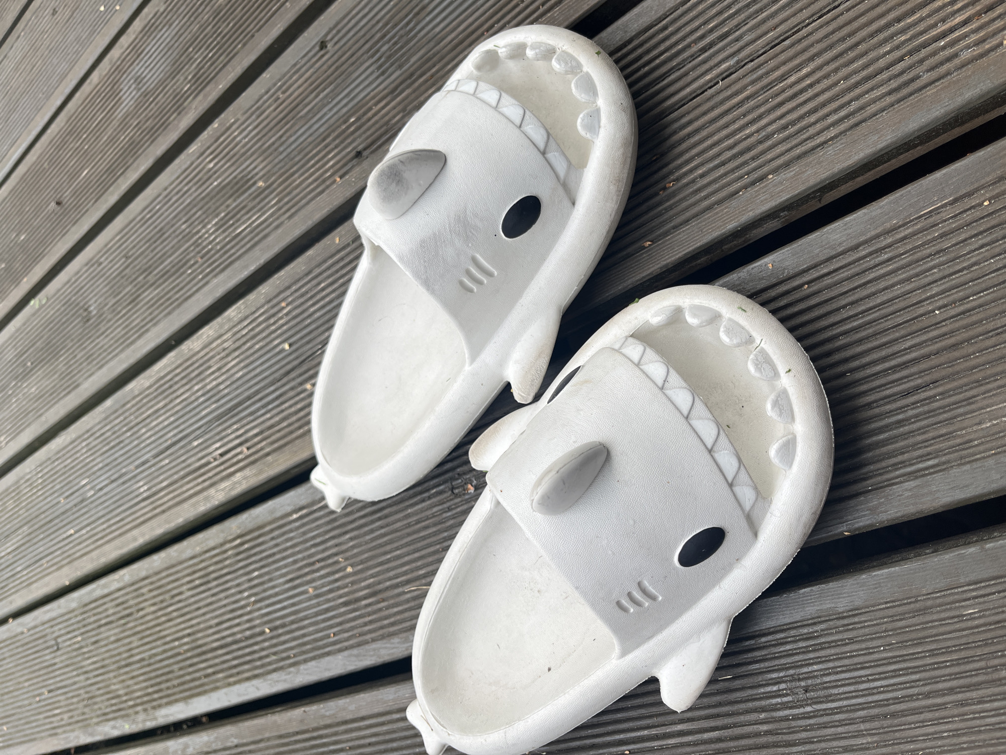 A pair of white shark-shaped slides are placed on a weathered, gray wooden deck. The slides are designed to resemble the head of a shark, with fins on the top, black circular eyes, and rows of white teeth along the open front. The deck appears to be outdoors, with visible gaps between the wooden planks and some scattered debris, possibly leaves. The scene is captured from a high-angle perspective, looking down at the slides. There are no people or animals present in the image. The lighting suggests it is daytime. This scene is located in Haabneeme alevik, Estonia.