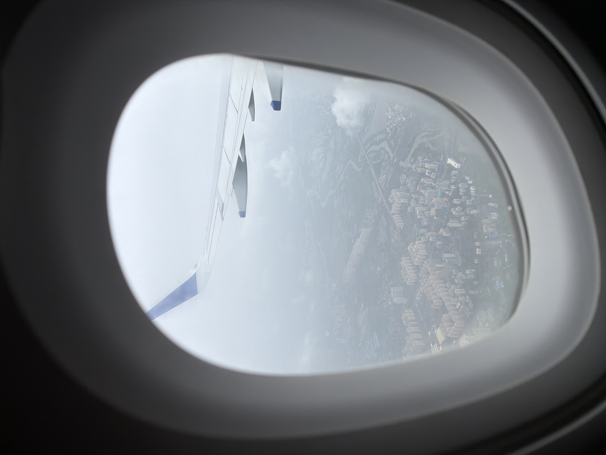An aerial view of a city is seen through an airplane window. The city is characterized by dense housing with many apartment buildings, some of which are light-colored and appear to be mid-rise. A winding river or waterway cuts through the urban landscape, with bridges visible crossing it. In the foreground, the wing of the airplane is visible, extending into the frame from the top left. The wing is white with blue accents on the wingtips. The overall atmosphere is hazy and overcast, suggesting a cloudy day or smog. The image appears to be taken from an aircraft at a moderate altitude, looking down on the city. The scene is consistent with an urban area, and the provided context indicates it is the city of Panvel, India.