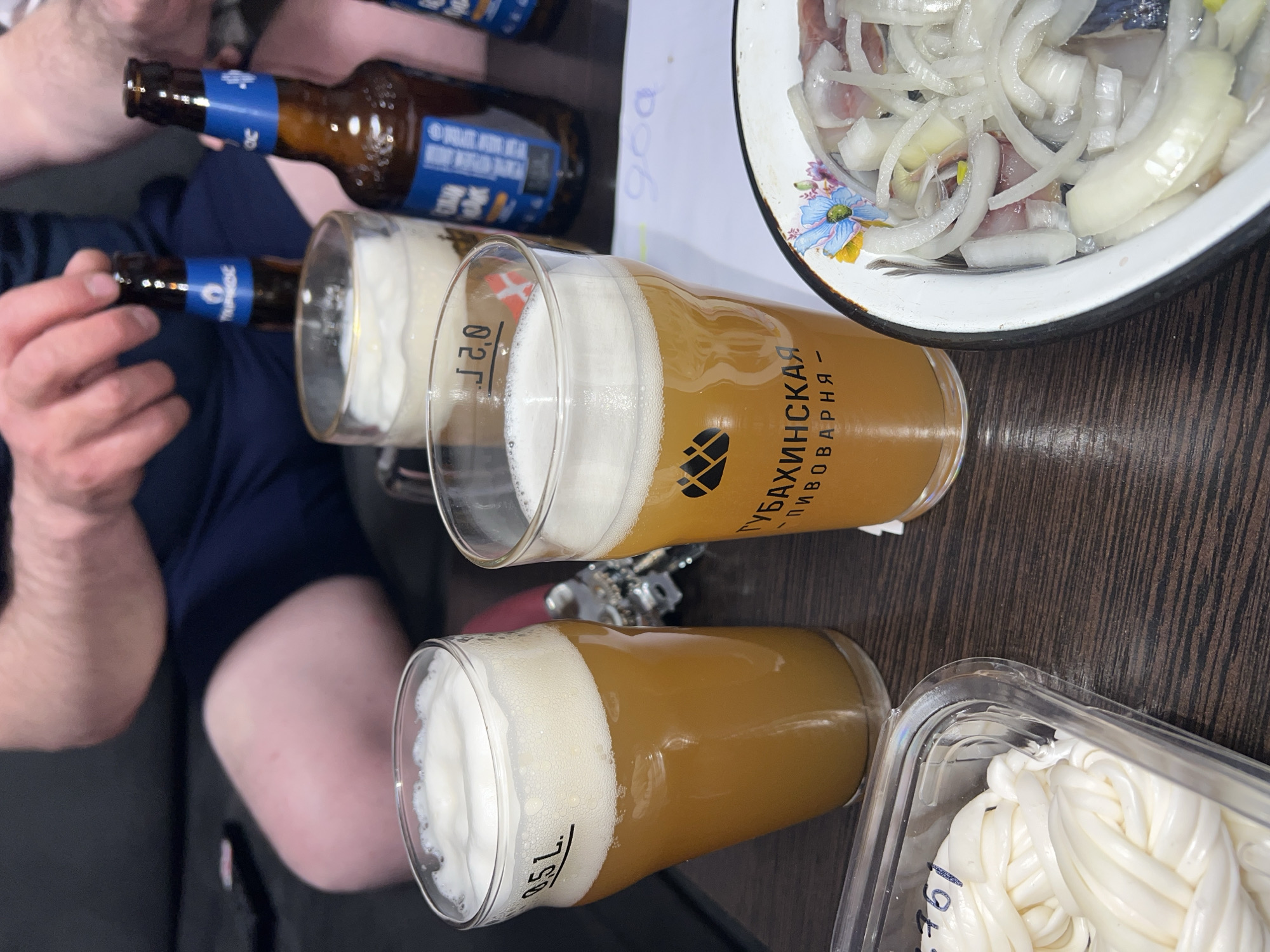 This image captures a casual indoor gathering, likely in Perm, Russia, featuring beer and traditional local snacks arranged on a dark wooden table.
The scene includes the arms and hands of at least two people. Three glasses of light-colored beer, each topped with a thick, frothy head, are visible. One glass has a "0.5 L" marking, while another indicates "0.2 L." A prominent beer glass clearly displays the Russian text "ГУБАХИНСКАЯ ПИВОВАРНЯ" (Gubakhinskaya Pivovarnya), identifying the brew as from a brewery in Gubakha, a town located within Russia's Perm Krai.
Accompanying the beer are two popular appetizers. A white enameled bowl with a blue floral pattern holds sliced herring generously topped with white raw onion rings. Beside the glasses, a clear plastic container contains braided white cheese, resembling varieties such as Suluguni or Chechil. The number "761" is handwritten on this container, possibly a price or product code.
Partially visible are two brown beer bottles with blue labels, one of which is held by a hand. Some text on the labels, though blurry, includes what appears to be "TUMEC" and parts of "ШИВОР" (likely "Живое," meaning 'live' or unpasteurized). In the background, a piece of white paper with the number "900" is also visible.
The overall impression is that of a relaxed social occasion, possibly an evening gathering, where individuals are enjoying local Russian beer paired with classic regional snacks.
