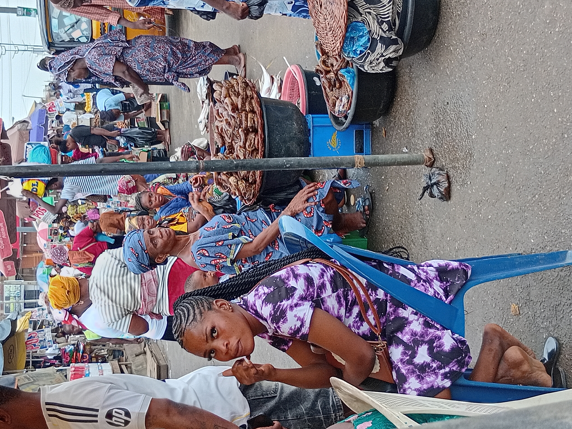 The image depicts a bustling outdoor market in Ikorodu, Nigeria. Numerous individuals, mostly women, are seen sitting and interacting with potential customers, while displaying their merchandise. The variety of goods include what appears to be dried fish in large buckets, clothing, and other general merchandise. The environment seems crowded and lively, with vendors set up along the street. The weather appears to be overcast or cloudy. The focus is on the vendors and the informal market atmosphere.