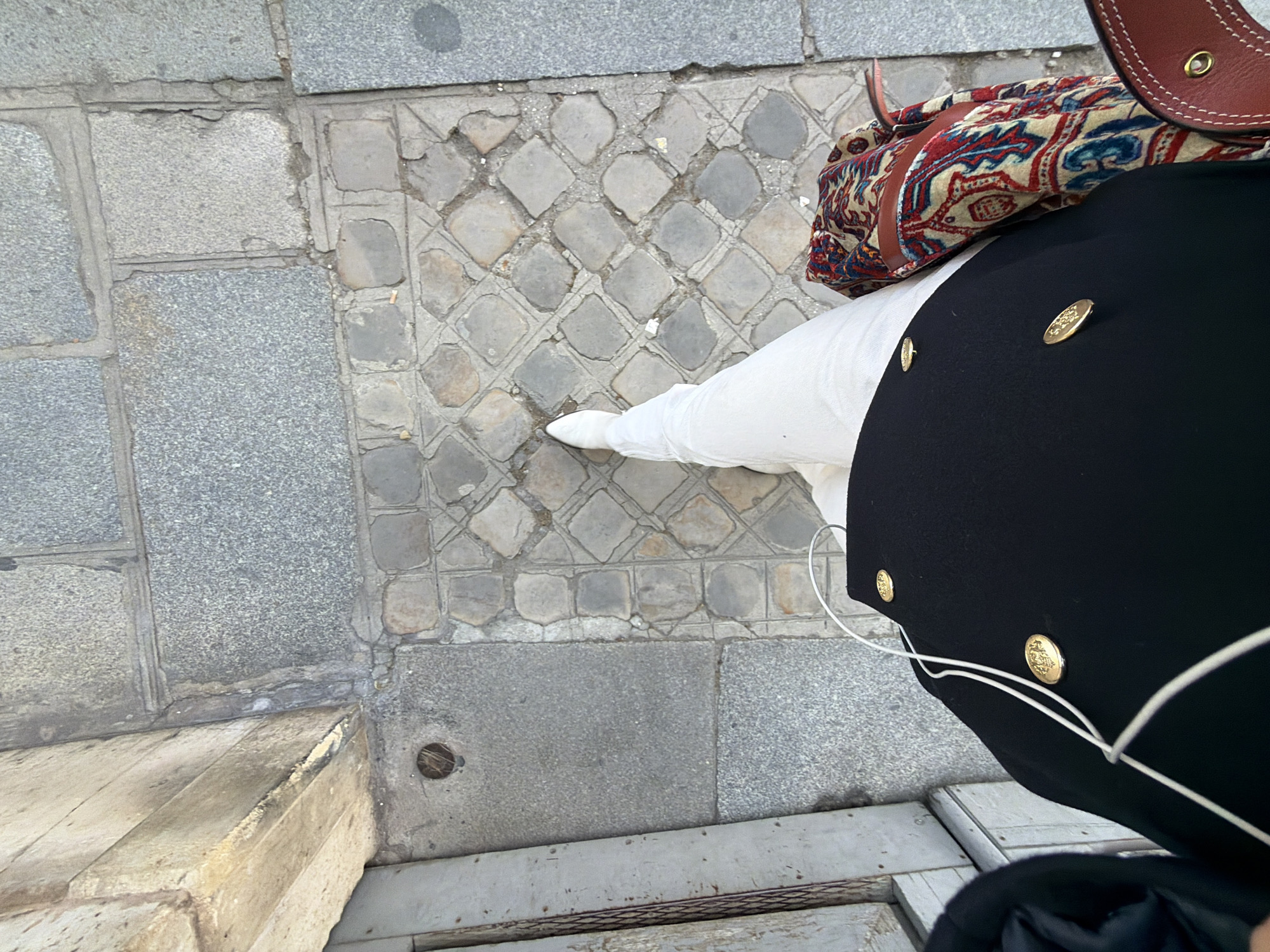 An overhead view captures a person's lower torso and legs standing on a city sidewalk in Paris, France. The individual wears a black garment, possibly a coat or jacket, featuring three visible round gold-colored buttons with embossed designs. White wired earphones drape across this black garment. Beneath, a white article of clothing, likely a shirt or trousers, is visible. The person's legs are clad in white trousers, with one foot extended forward, wearing a white pointed shoe or boot. To the person's right, a patterned textile item, possibly a scarf or part of a bag, displays a beige background with red, blue, and black intricate motifs. Adjacent to this, a section of a brown leather object, featuring prominent stitching and a brass eyelet, is visible, consistent with a bag strap.
The ground surface consists of two distinct paving types. To the left and upper right, larger, rectangular grey granite paving stones are present, some exhibiting minor scuffs and wear. Adjacent to these, particularly in the center and towards the lower right, are smaller, irregularly shaped grey and tan cobblestones arranged in a repeating diamond or basket-weave pattern. This cobblestone section shows signs of age, including worn edges and some gaps between stones. Near the left edge of the frame, where the granite paving meets another surface, a small circular metal or concrete drain is visible. A dark, weathered wooden or metal structure, possibly a door frame or gridded grate, is partially visible along the far left edge of the scene. Minor debris, including a cigarette butt fragment, is scattered on the cobblestones.