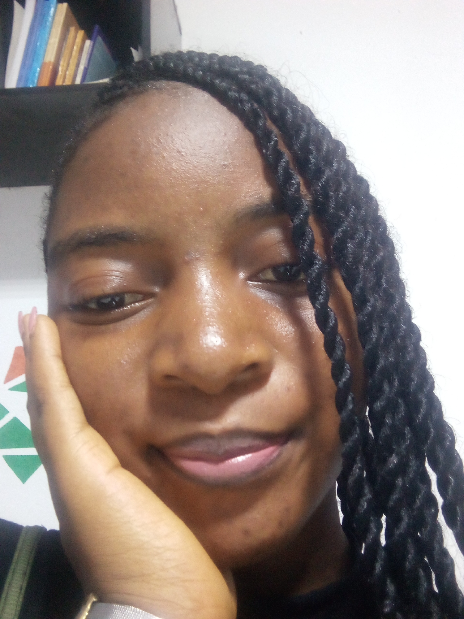 A medium shot of a Black woman is shown from the chest up, as she holds her hand to her face and smiles. She has dark skin, and her dark hair is braided into long twists. She is looking at the camera and her mouth is slightly open, revealing her teeth. The background appears to be indoors, with a shelf of books visible on the left side of the image and a white wall behind her. The lighting is bright and direct, creating highlights on her skin. The overall impression is that of a casual, informal photograph.