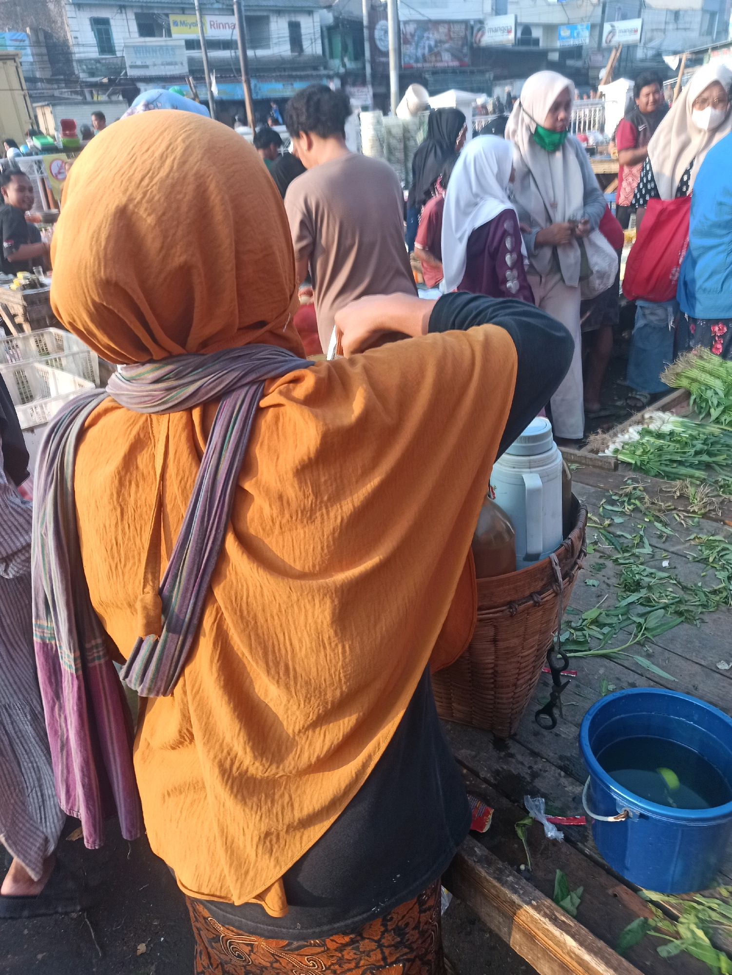 The image depicts a bustling market scene in Cikarang, Indonesia, likely near the city center or a prominent market area. In the foreground, a person wearing an orange headscarf and a traditional patterned skirt is partially visible from behind, facing away from the camera. They are also wearing a striped scarf draped around their neck. To their right, a woven basket contains a thermos and a bottle. A blue bucket filled with water and a slice of lime sits on a wooden platform in the lower right corner.
The background reveals a lively market environment with numerous people engaged in various activities. Several individuals are wearing hijabs and traditional Indonesian clothing, suggesting a strong cultural presence. Stalls are set up, some displaying fresh produce like green onions. Buildings and signs are visible in the distance, indicating an urban setting. The overall atmosphere suggests a typical day in a vibrant Indonesian marketplace.
