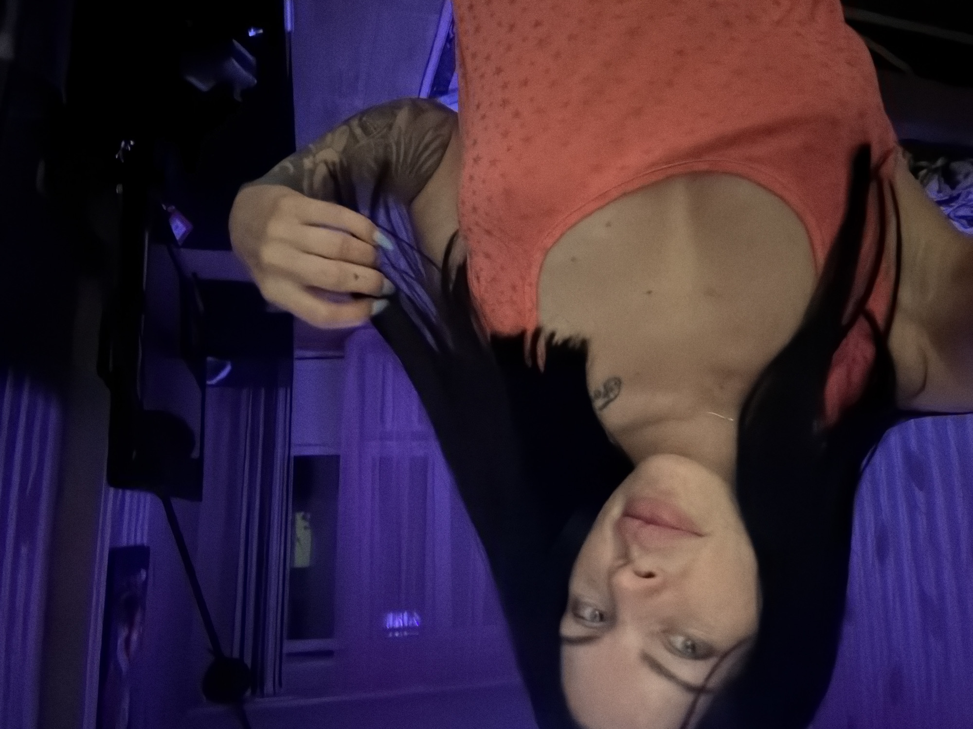 The image is a medium shot of a woman with dark hair and fair skin, wearing a coral-colored tank top. She is looking towards the camera with a neutral expression. Her left hand is raised, holding some of her hair. Tattoos are visible on her right arm and shoulder. The background is dimly lit with purple lighting, with vertical striped curtains and a window visible. A dark, unidentifiable object is on the right side of the frame, possibly a piece of furniture or equipment. The scene appears to be indoors, in a room with low lighting and a vibrant purple hue, suggesting a setting that could be a bedroom or a living space with mood lighting. No specific location in Ufa, Russia, can be identified from the image.