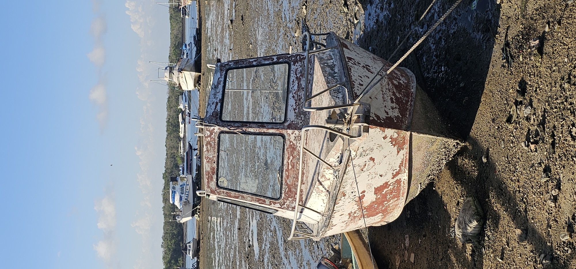 The image depicts a muddy landscape at low tide in an area with several boats, potentially a harbor or riverbank. The foreground features a derelict boat cabin, its paint peeling and rusting. The cabin has two rectangular windows and metal railings. A thick rope is tied to the boat cabin, extending into the background. The background shows other boats moored in the mudflats at low tide. The sky is blue with scattered clouds. It's not possible to verify the location or the city of Unknown, Indonesia.
