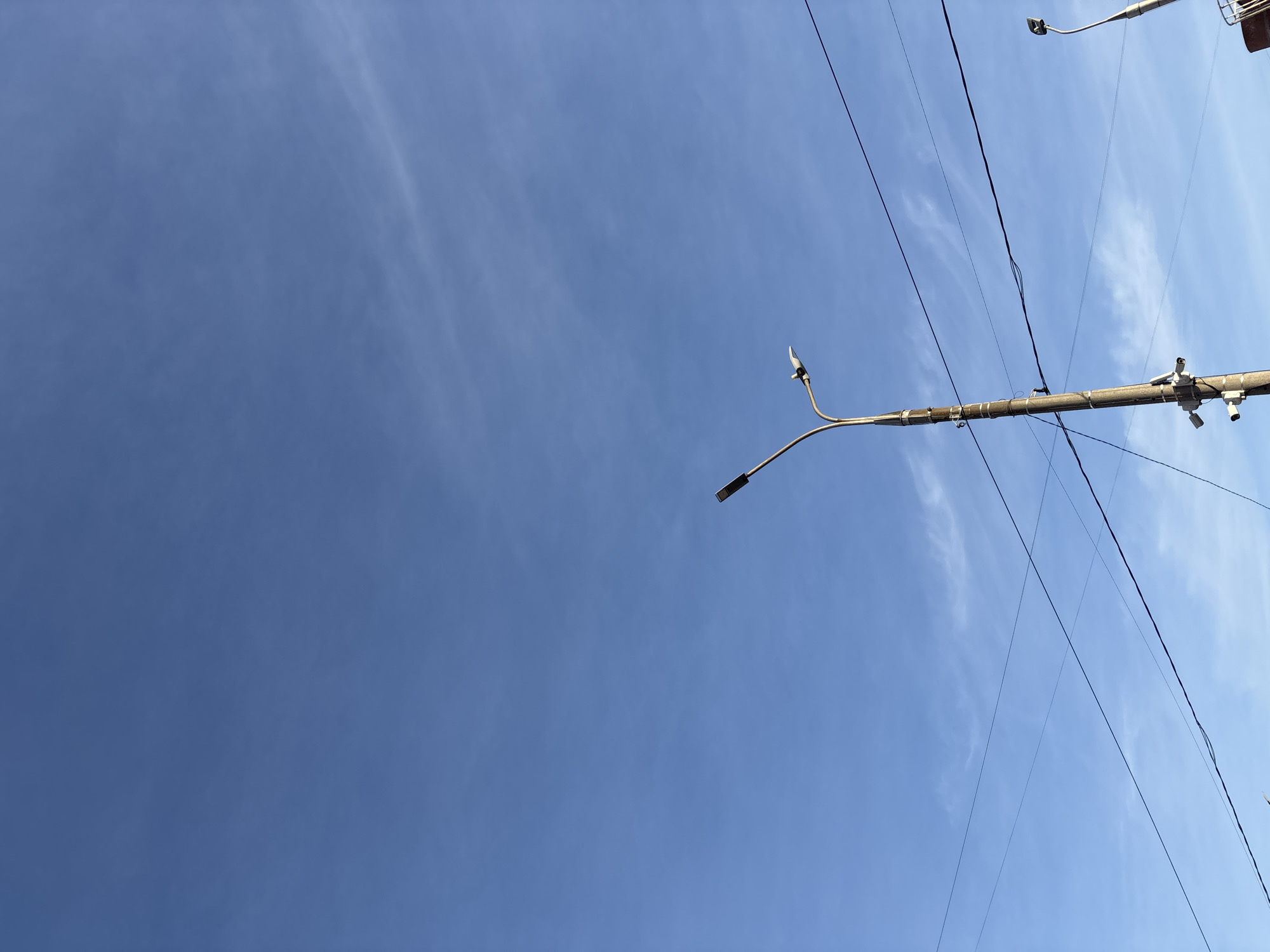 The image depicts a low-angle view of a clear blue sky with sparse cirrus clouds. Several power lines traverse the upper portion of the frame. There is a street light pole mounted on a utility pole with two streetlight heads. Additionally, three security cameras are attached to the utility pole. The location of the scene is Perm, Russia.