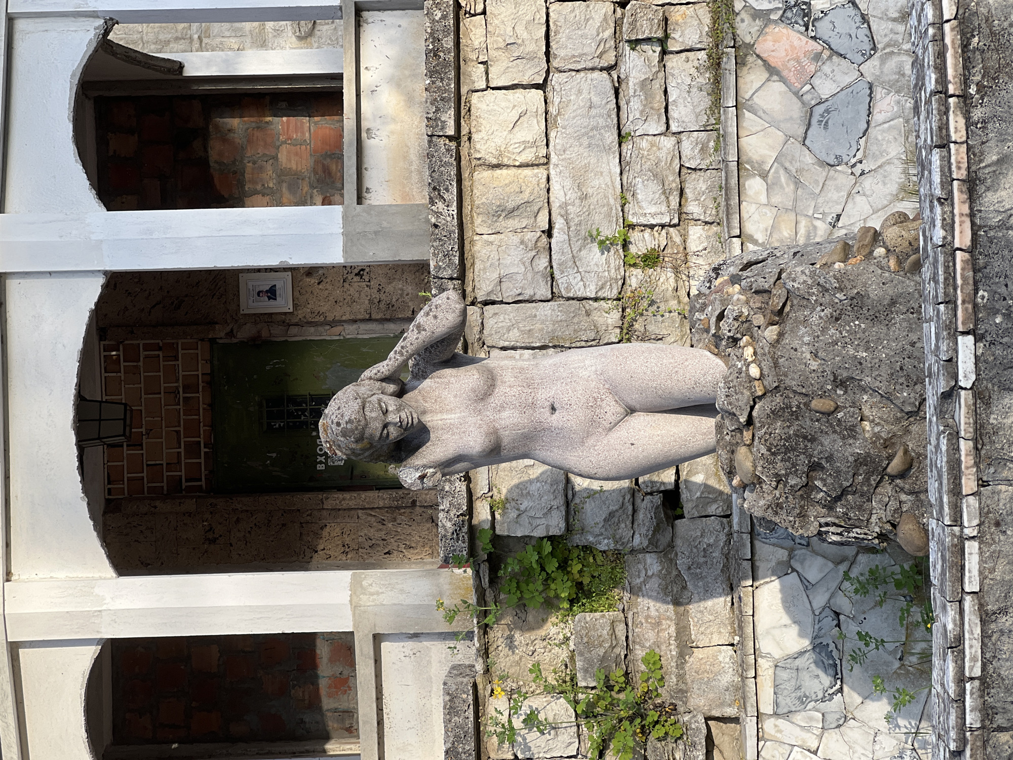 The image, oriented correctly, depicts an outdoor scene featuring a reclining nude female stone statue set amidst various architectural elements. The light-colored statue, with one arm behind her head, rests on a rough stone base. To her left, a multi-tiered structure with white, perhaps concrete, frames contains arched openings revealing red brick walls within. Inside one of these openings, a small framed photograph of a man in a dark suit is visible on a wall. Below this, a dark green surface, possibly a door, shows faint Cyrillic text, including the word "ВХОД" (Entrance).
The ground and walls around the statue are made of different types of stone. A wall of rough-hewn light grey blocks stands behind the statue, while to her right, an irregular mosaic of stone slabs forms a path or ground cover. A dark, irregular rock formation containing smooth, rounded pebbles is also present. Small green plants and weeds sprout from cracks and crevices in the stonework. The scene is bathed in bright daylight, casting distinct shadows and suggesting a clear day. The architectural style, the statue, and the framed photograph hint at a memorial, mausoleum, or an artistic garden installation. The white structural elements show some signs of weathering or an unfinished appearance with jagged edges.
