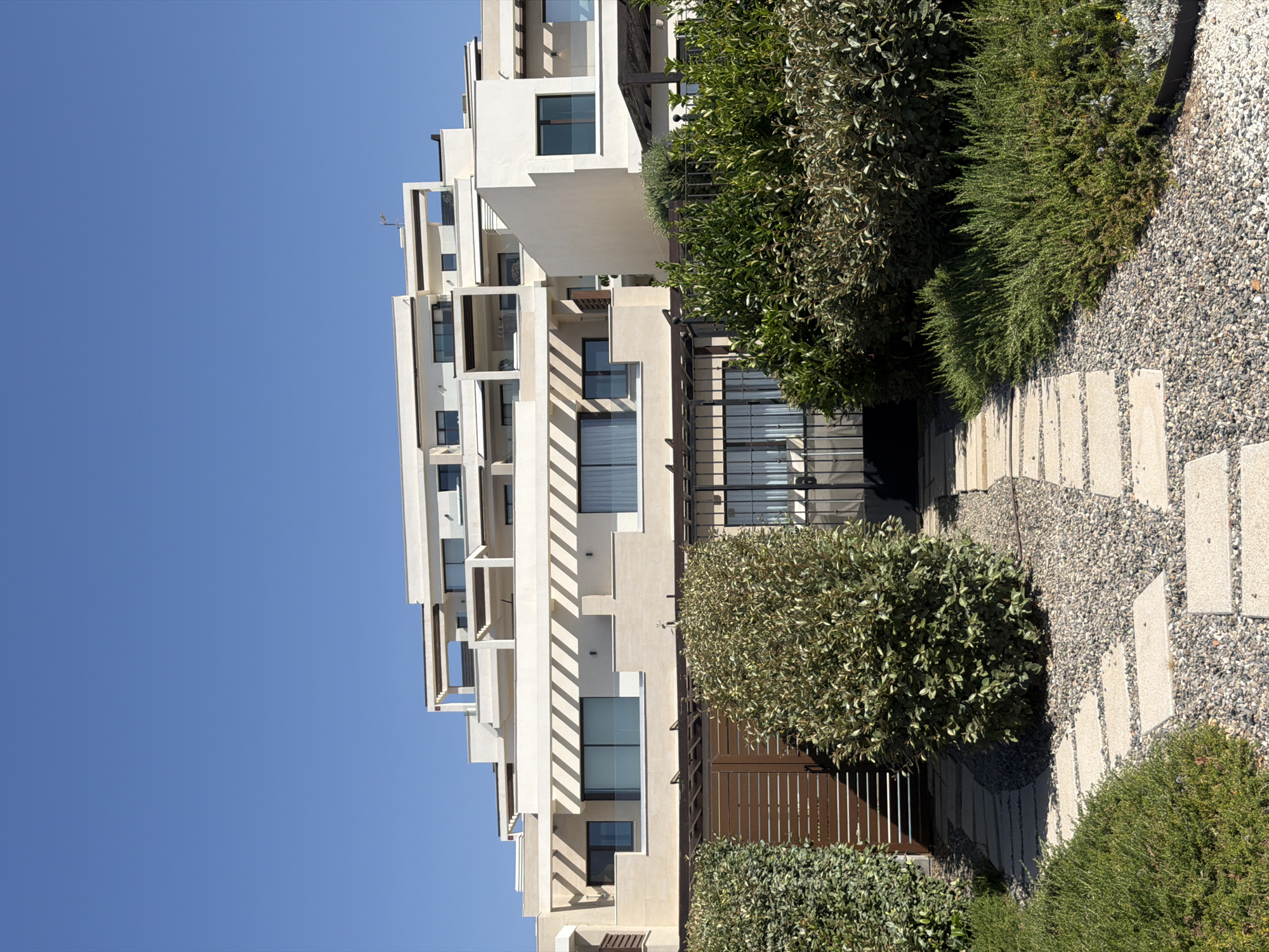 The image shows a contemporary multi-level residential building with light-colored facade and a clear blue sky. The building features several balconies and terraces with pergolas. In the foreground, there is a pathway made of large, square stone slabs laid on a bed of gravel. Flanking the pathway are lush green bushes and trees. A brown slatted gate and a black metal fence are visible, separating the garden area from the building's entrance. The scene appears to be a residential complex in Estepona, Spain, under bright daylight conditions. There are no people visible in the image.