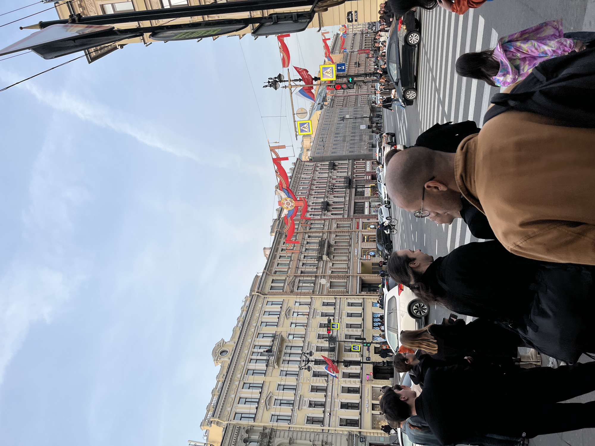 The image shows a street scene in an unidentified city with buildings adorned with flags. Pedestrians are present, and vehicles are visible in the street. The sky is partially visible with clouds. The general environment is urban, appearing to be a city street with pedestrians and vehicular traffic.