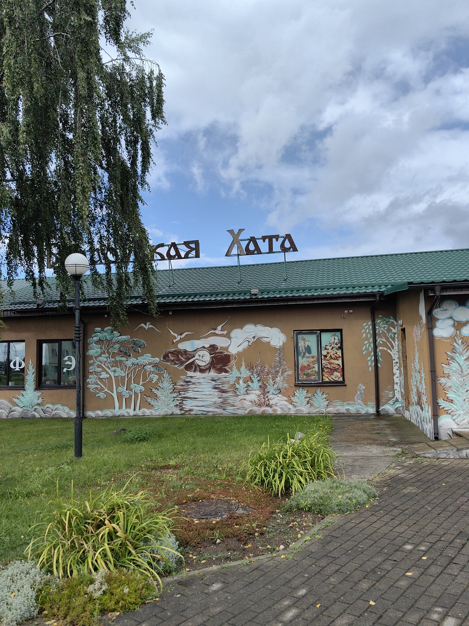 The image shows the facade of a building with a mural. The building is located in Myadzyel, Belarus. The facade is tan, and the roof is green. The mural depicts a landscape with trees, birds, and a cloudy sky with a face. The building has several windows, and there is a sign on the roof that reads "КАЯ ХАТА". In front of the building, there is a lamp post, a lawn, and a paved walkway. A large tree with drooping branches is on the left side of the image.