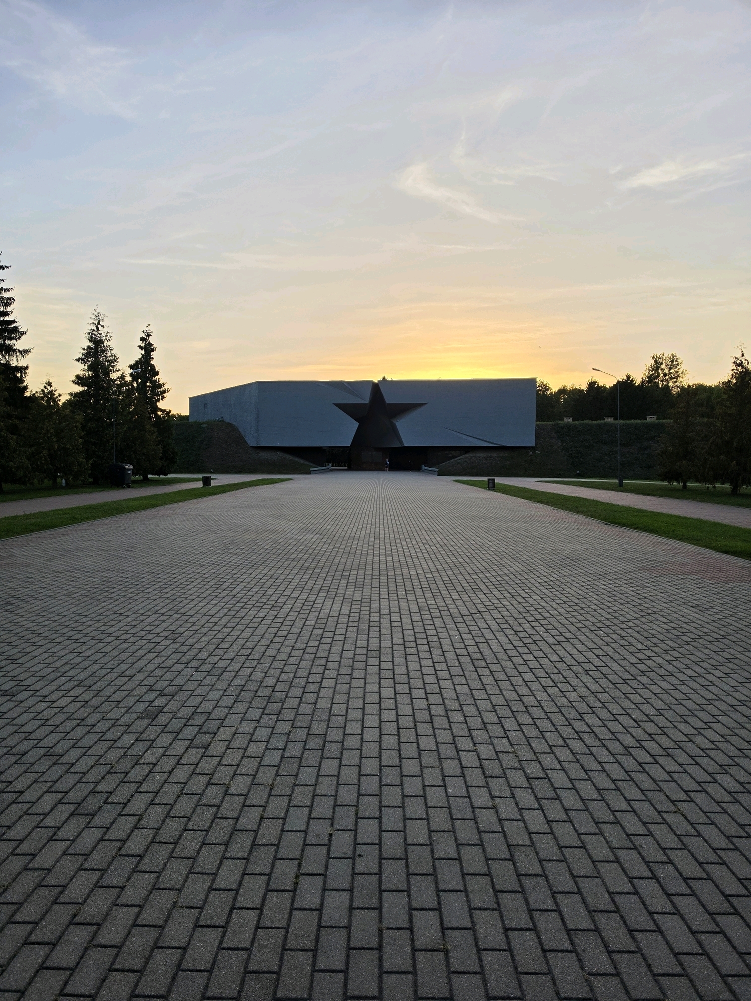 The image shows an exterior view of the Brest Fortress Memorial Complex in Brest, Belarus, at dusk. A wide, paved walkway made of rectangular grey bricks leads towards a monumental building with a large, dark star-shaped structure embedded in its facade. The building is a light grey color and appears to be constructed from concrete or a similar material, with sharp, angular lines. The sky above is a soft gradient of pale blue and yellow, indicating the setting sun. On either side of the walkway, there are strips of green grass, followed by trees and a raised grassy embankment leading up to the fortress. A lamppost is visible to the right of the walkway, and a couple of dark trash receptacles are also present. The overall scene is serene and somber, characteristic of a memorial site. There are no people clearly visible in the main part of the image, though a small figure might be present near the entrance of the building on the left.