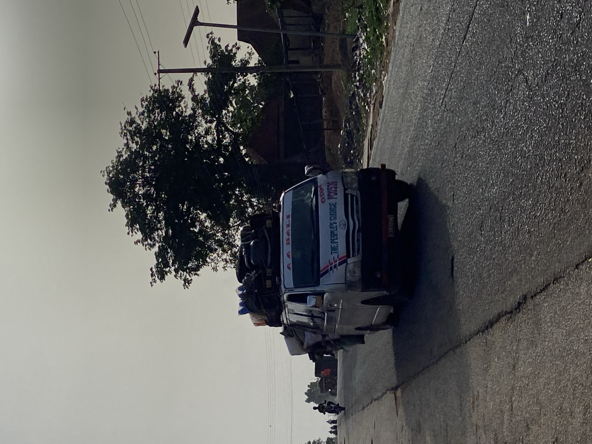 This image, captured in Mararaba, Nigeria, depicts a daytime street scene featuring a heavily loaded white commercial mini-van traveling on an asphalt road. The sky appears bright but overcast or hazy, with power lines and utility poles visible against its greyish expanse. A large tree with dense, dark green foliage stands prominently to the left of the road.
The mini-van is a central subject, packed with various items and luggage secured to its roof rack, indicating its use for transporting goods or passengers. Visible text on the front of the vehicle includes "AA BALI" and "THE PEOPLE'S CHOICE". In the background, behind the tree and to the side of the road, there are indistinct structures or buildings and an area of unkempt ground with some visible debris. Further down the road, smaller figures, likely people or other vehicles, can be discerned, suggesting ongoing daily activity. The scene captures a common sight of roadside transport and infrastructure in the region.