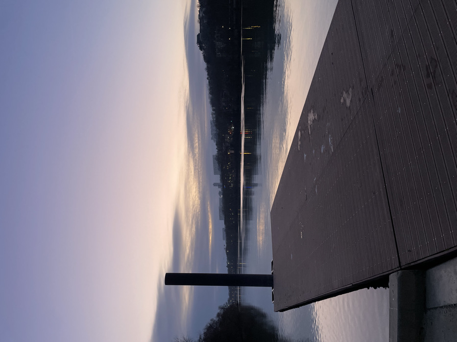 A lakeside dusk scene displays a wooden dock extending into still water. A dark post anchors the dock. Across the water, faint city lights glimmer against a backdrop of shadowy hills. The sky above is a blend of pale purples and yellows, reflecting in the water's surface. The dock's planked texture and the smooth water contrast the natural, silhouetted landscape.