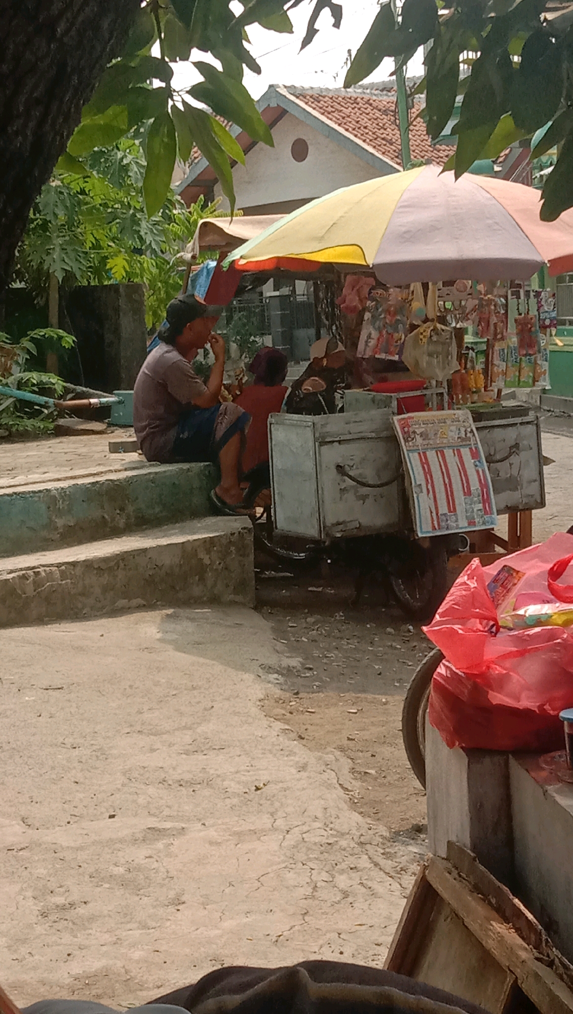 In Ngempit, Indonesia, a street vendor's cart sits under a colorful umbrella. The cart is equipped with two large metal boxes, likely for storing goods, and a smaller box in front displaying a printed chart with rows of red and white stripes and smaller images. Bags of snacks and other items are displayed on the cart's shelves. A person in a brown shirt and blue patterned shorts sits on concrete steps to the left of the cart, facing it and appearing to be engaged in eating or smoking. Behind them, another person in a red shirt is partially visible. The ground around the cart is dusty and uneven. In the foreground, a large red plastic bag is visible, and to the far right, a wooden frame lies on the ground. The scene is partially shaded by tree leaves in the upper portion of the image. A residential building with a tiled roof is visible in the background.