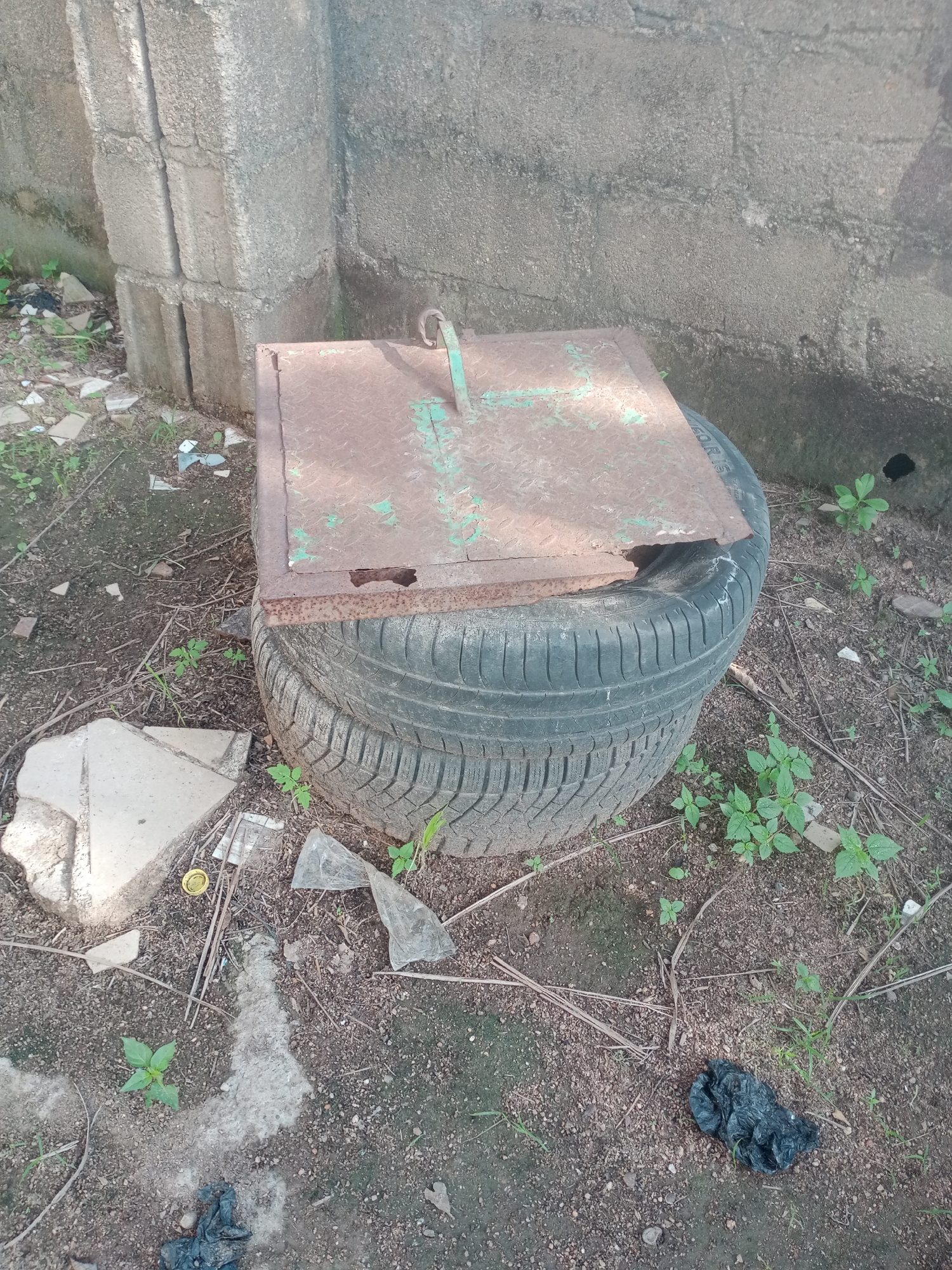 In Jalingo, Nigeria, an outdoor scene shows two stacked tires supporting a rusty metal cover with a green handle. The tires sit on the ground next to a textured concrete wall and some stacked concrete blocks. The ground is covered with soil, small stones, dried leaves, and scattered debris, including pieces of broken concrete, plastic fragments, and a yellow bottle cap. Several small green plants are growing in the soil around the tires and debris. To the right of the tires, a pile of black, crumpled material, possibly trash or animal waste, rests on the ground.