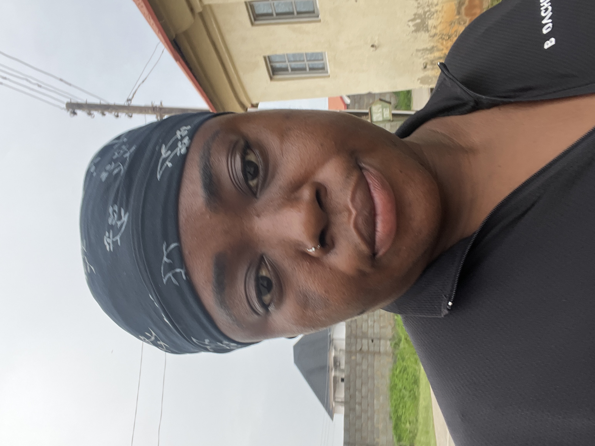 A close-up selfie depicts a dark-skinned individual with a nose piercing, wearing a black patterned headscarf and a black zip-up athletic shirt. The background reveals elements of an urban environment, including an overcast sky, utility poles and wires, a beige two-story building with two visible windows, a brick fence, and a section of lawn. The location is identified as Abuja, Nigeria.