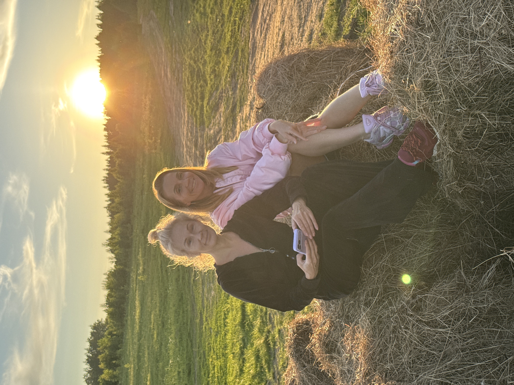 The image displays two women seated outdoors during sunset in a rural or pastoral setting, identified as Стеклянный, Russia. The photograph is oriented as if the sun is on the left, casting a strong golden backlight across the scene.
The setting sun is visible on the left side of the frame, low on the horizon behind a line of trees, creating a bright glow and warm light. The sky has some wispy clouds. The ground where the women are sitting is covered in dry grass or hay, with a noticeable hay bale partly visible behind one of them. In the background, a field of green grass or crops stretches towards the tree line.
One woman, with light brown hair, is wearing a light pink long-sleeved jacket or top. She smiles warmly at the camera, her legs extended, wearing light-colored athletic shoes (one purple, one reddish-purple). She appears to be leaning against or sitting on a mound of hay.
The second woman, with blonde hair tied back, is wearing a dark, possibly black, long-sleeved zipped top and dark pants. She is also smiling at the camera, holding a light-colored smartphone in her hands. A small, glowing yellow-green object is discernible on the dry grass near her leg.
The women are engaged in the activity of posing for a photograph. Their smiles convey a relaxed and joyful emotion. The time of day is clearly sunset, characterized by the low sun and golden hour lighting. No visible text is present in the image.
