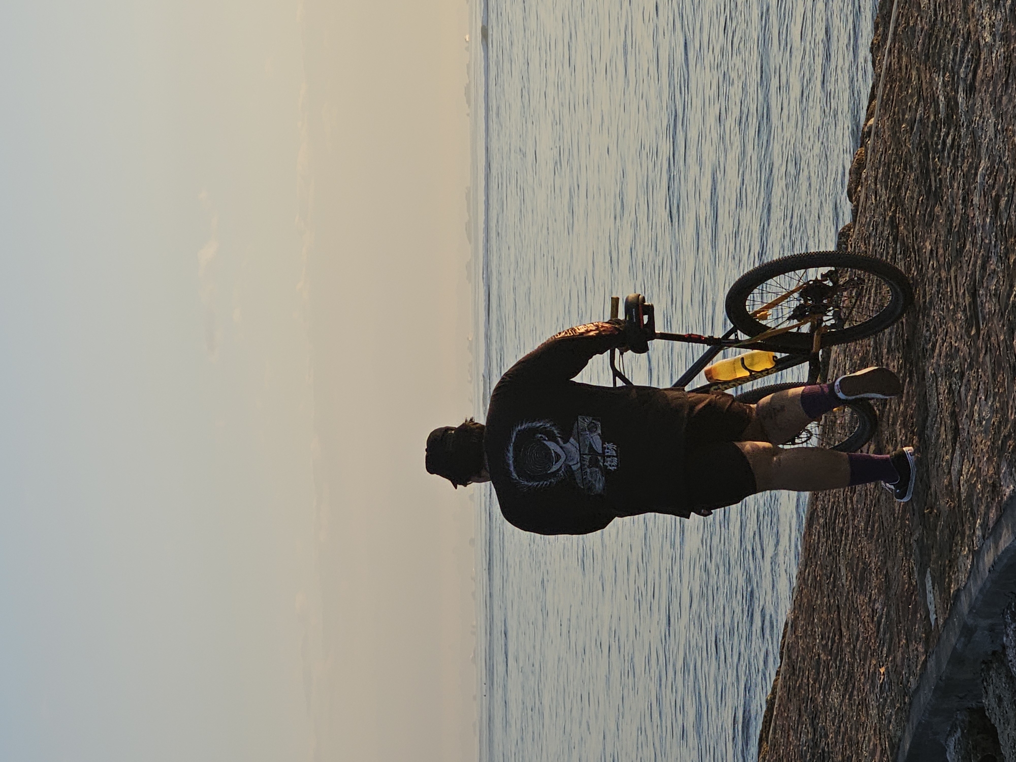 A person wearing a black t-shirt with a graphic print, black shorts, a black cap, purple socks, and black and white sneakers walks along a stone embankment next to a body of water. The person is pushing a bicycle with a yellow water bottle attached to its frame. The bicycle has black tires and yellow accents on the frame. The water is calm and reflects the soft light of what appears to be early morning or late afternoon. In the distance, there are several small boats or buoys visible on the horizon. The sky above is a pale blue with hints of yellow and orange, suggesting sunrise or sunset. The embankment is made of rough, uneven stones. The scene is likely located in a coastal area of Denpasar, Indonesia, possibly along a beach promenade or a seaside pathway.
