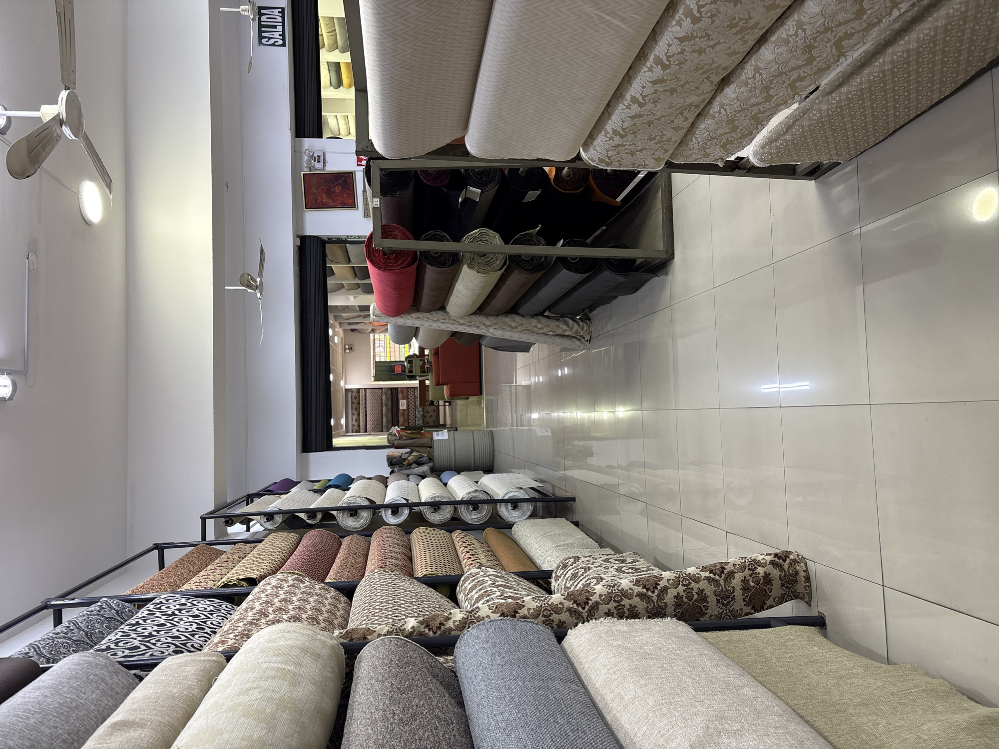 The image depicts the interior of a fabric store in La Victoria, Peru. The store is filled with large rolls of fabric displayed on metal shelving units that line the aisles. The fabrics vary in color, pattern, and texture, ranging from neutral tones like beige and grey to more vibrant hues like red and purple. Some rolls are plain, while others feature intricate floral or geometric designs. In the background, a sign reads "SALIDA," indicating an exit. Two ceiling fans are visible, suggesting the environment is warm. The floor is tiled with a reflective surface, creating a polished appearance. The overall scene is one of an organized retail space dedicated to the sale of textiles.