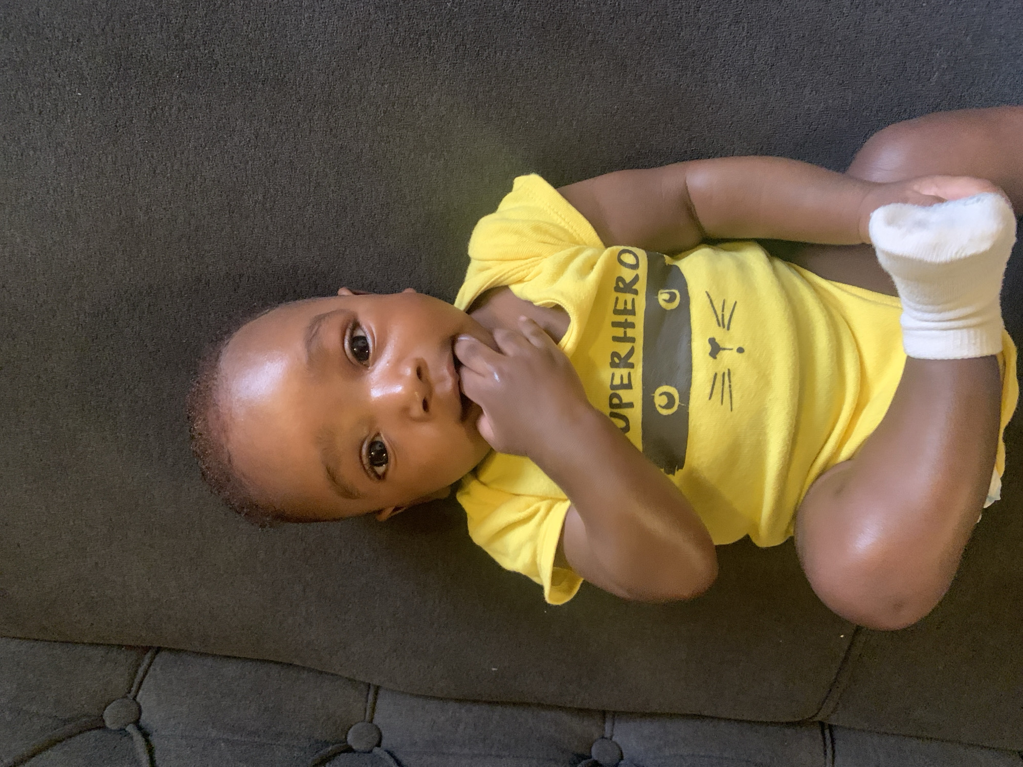 A baby lies on a dark grey, tufted sofa. The baby is wearing a yellow onesie with a cartoon cat face and the word "SUPERHERO" on it. The baby has dark skin and is wearing a white sock on one foot. The baby's hand is in its mouth, and it is looking directly at the camera. The location is unknown and there is no reference to the city of Barakin Akawo, Nigeria.