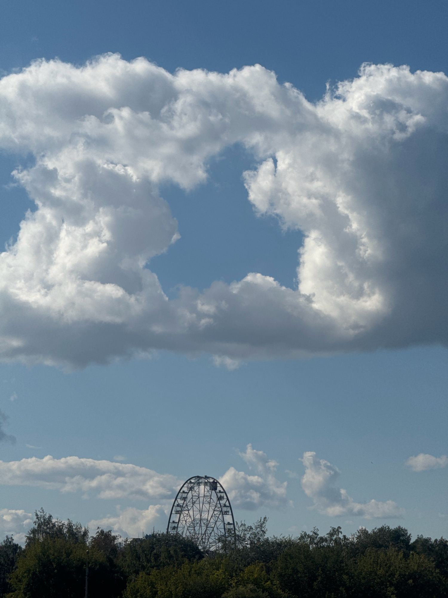 The image depicts a Ferris wheel silhouetted against a blue sky filled with dramatic, fluffy white clouds. The Ferris wheel is located behind a dense line of dark green trees, forming a horizon. The clouds are concentrated in the upper portion of the frame, with a large, somewhat amorphous gap in the center revealing more of the sky. The lower portion of the image shows the tops of the trees and the wheel. No people are visible. The scene suggests an outdoor recreational area or park. The location is identified as Irkutsk, Russia.