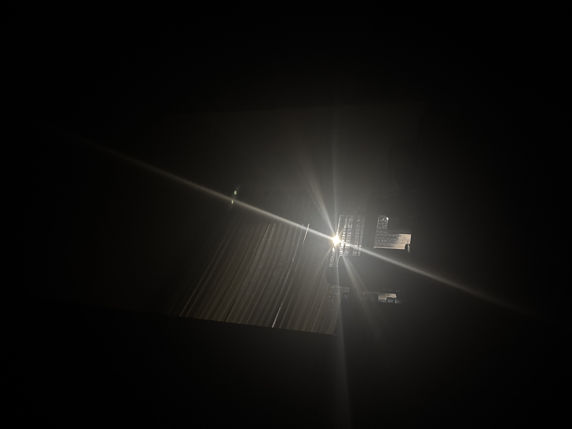 The image depicts a nighttime scene in Welshpool, United Kingdom. Light emanates from what appears to be a building with corrugated metal siding. The light source is bright, causing a lens flare effect. The environment is dark, obscuring details outside of the immediate area illuminated by the light. There are no visible people or interactions in the image.