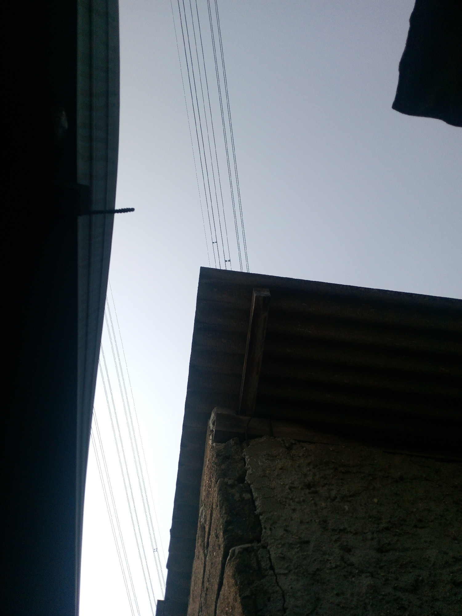 The image shows a low-angle view of overhead wires against a pale sky. On the left, a dark, curved structure, possibly a bridge or overpass, partially obscures the view. To the right, a corrugated metal roof on a rough concrete structure is visible. The scene appears to be in an urban or semi-urban environment, possibly Dukuh, Indonesia, given the context. There are no people or discernible actions depicted. The lighting suggests it is either early morning or late afternoon.
