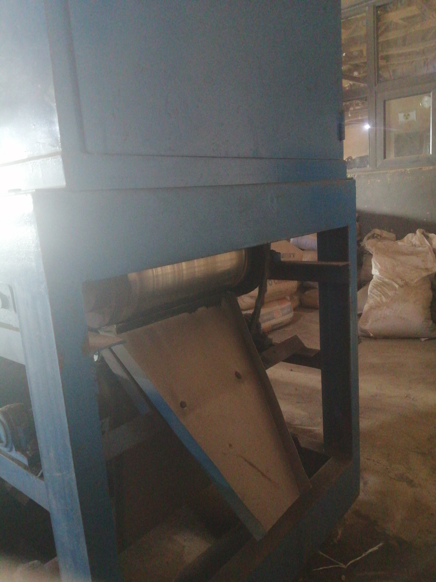 The image shows a close-up, eye-level, interior shot of industrial machinery and stacked bags in what appears to be a warehouse or factory. The main subject is a large, blue metal machine with visible internal components including a roller and a funnel-shaped chute made of light brown material. The machine is supported by a dark blue metal frame. To the right of the machine, several large, off-white sacks are piled up. In the background, a window with multiple panes is visible, revealing the interior of another part of the building with some wooden structures and possibly more sacks. The floor is concrete and appears somewhat worn. The lighting is dim, with a strong light source coming from the left, creating reflections on the machine's surface. There are no people or active processes depicted. The scene is static and gives the impression of an operational or storage area within an industrial facility.