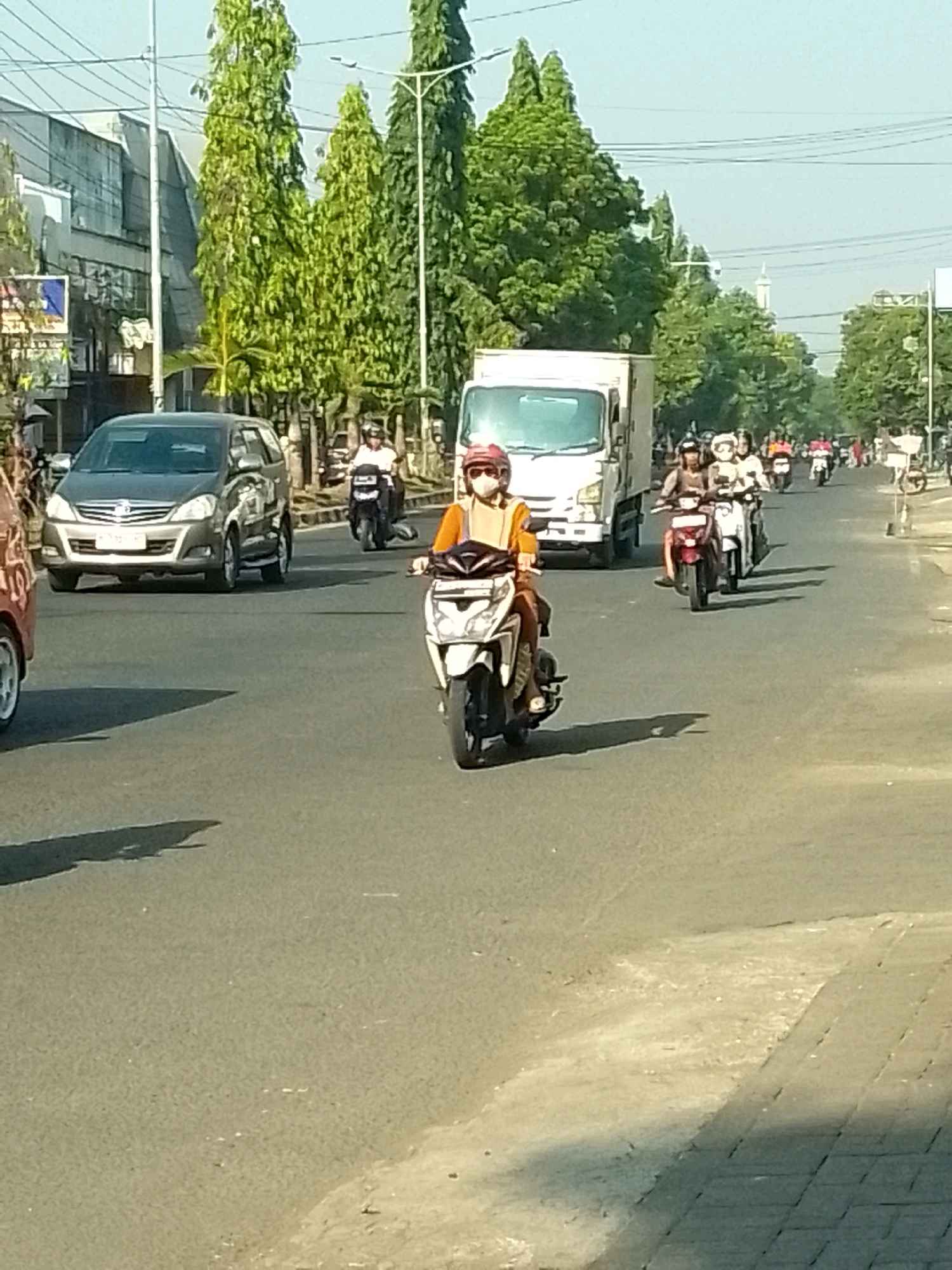 The image depicts a street scene in Jember, Indonesia, with several motorcycles and a truck on the road. In the foreground, a person wearing a helmet and an orange shirt is riding a white scooter. A brown SUV is visible on the left side of the road. Further down the road, more motorcycles and a white truck are present. The environment appears to be a sunny day with trees lining the street and buildings in the background. The overall scene is one of moderate traffic flow on a paved urban road.