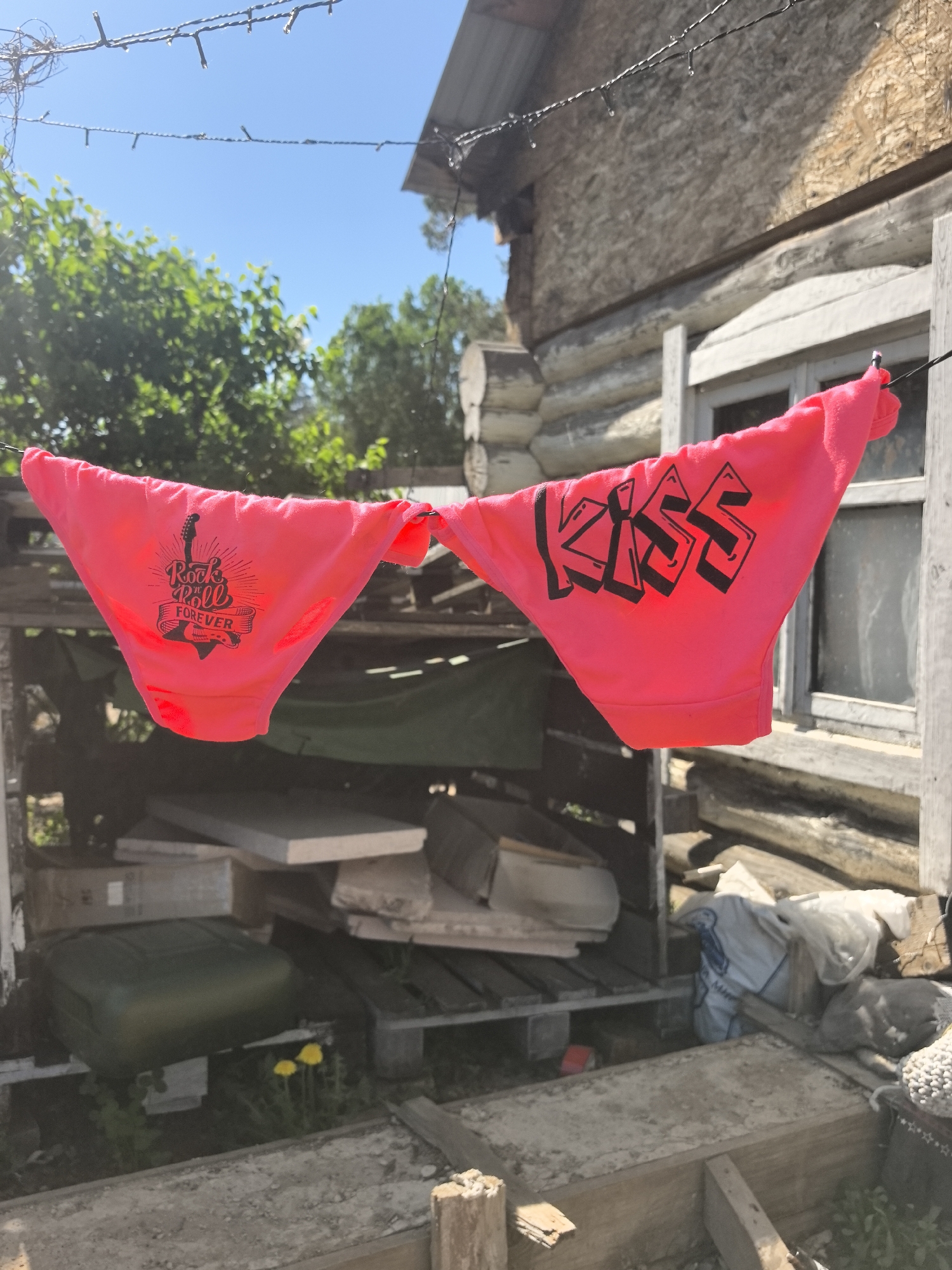 Two pairs of bright pink underwear are hanging on a clothesline. The pair on the left has "Rock 'n Roll FOREVER" printed on it, while the pair on the right has "KISS" printed on it. They are hanging in front of a cluttered wooden shed with various items, including a green container and white boards. The shed is located near a weathered, wooden house with a visible window frame. The sky is blue with some trees visible in the background. The scene appears to be in an outdoor area, possibly a backyard. There are no people visible in the image. No data suggests the image was taken in Noviy, Russia.