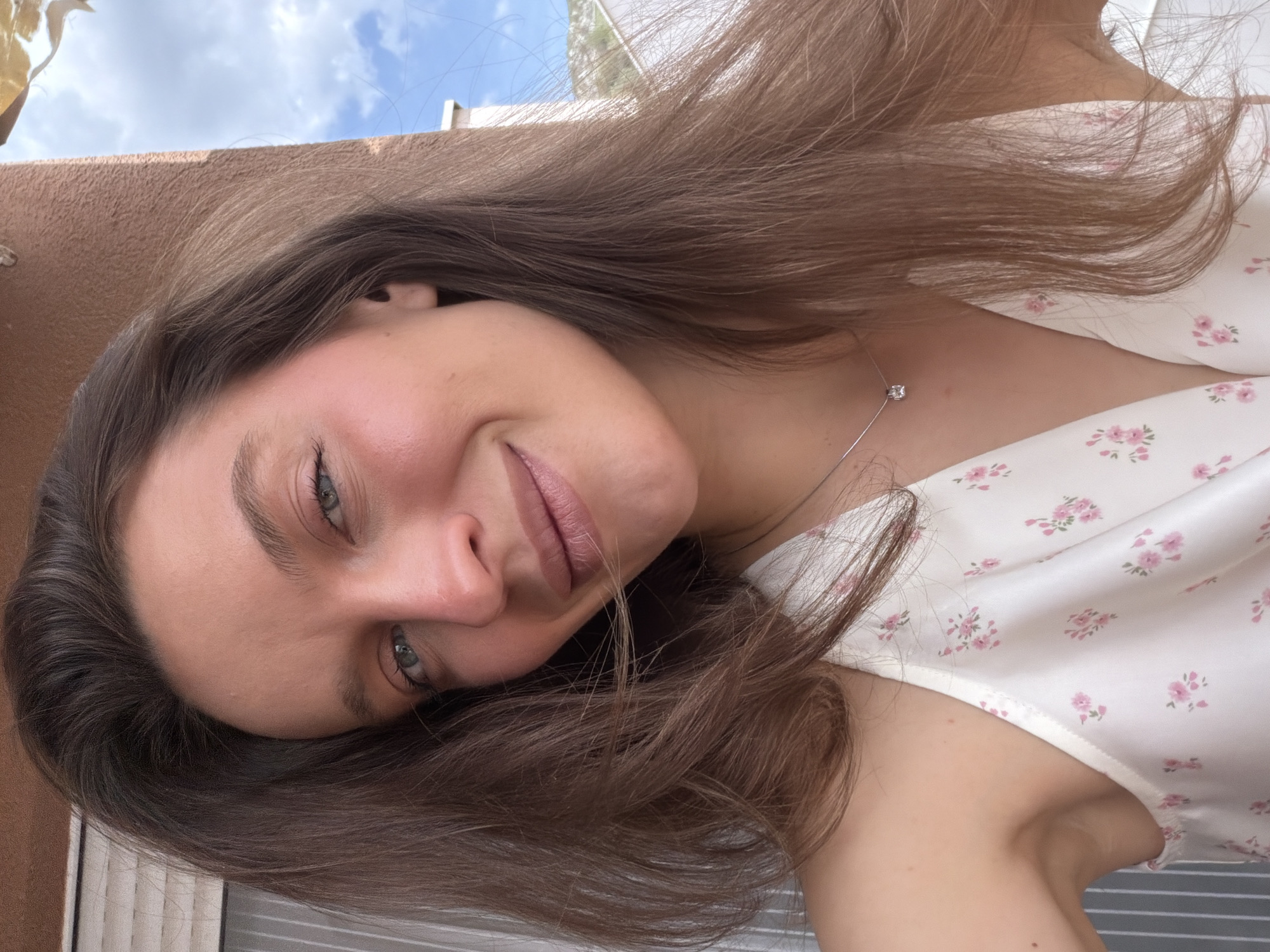 A young woman with long, slightly wavy brown hair is depicted in a close-up, upward-angled shot, likely a selfie, taken outdoors in Budva, Montenegro. Her head is tilted to the right as she gazes directly at the camera with a soft, contented smile. She has light-colored eyes, appearing blue or green, and naturally defined eyebrows. Her lips are a soft pinkish-brown, and a subtle flush is visible on her cheeks.
She is wearing a white top with a delicate pink floral pattern. A slender silver necklace featuring a small, clear, square-cut gem hangs around her neck. The background reveals a clear blue sky with scattered white clouds, positioned above a light brown or beige wall-like structure, suggesting an outdoor setting such as a balcony or terrace. The lighting indicates it is daytime, with natural light illuminating her features. The overall mood is calm and serene. No text is visible in the image.