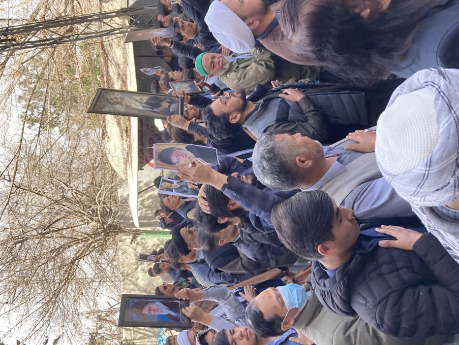 This image depicts a dense outdoor gathering of men in Quetta City Tehsil, Pakistan. The crowd consists primarily of adult males, many dressed in casual attire including jackets, vests, and traditional headwear such as turbans and kufis. Numerous individuals are seen with their right hand placed over their chest or heart, a gesture of respect or mourning. Several participants are holding aloft framed portraits and printed pictures; these include prominent images of Ayatollah Ali Khamenei and Ayatollah Ruhollah Khomeini, both identifiable by their black turbans and distinct features. Additionally, at least one taller framed image displays a classical-style depiction of a bearded figure. In the foreground, one man wears a light blue surgical mask. The background features bare branches of deciduous trees, suggesting a non-summer season, and a light-colored structure or stall with indistinct text visible.