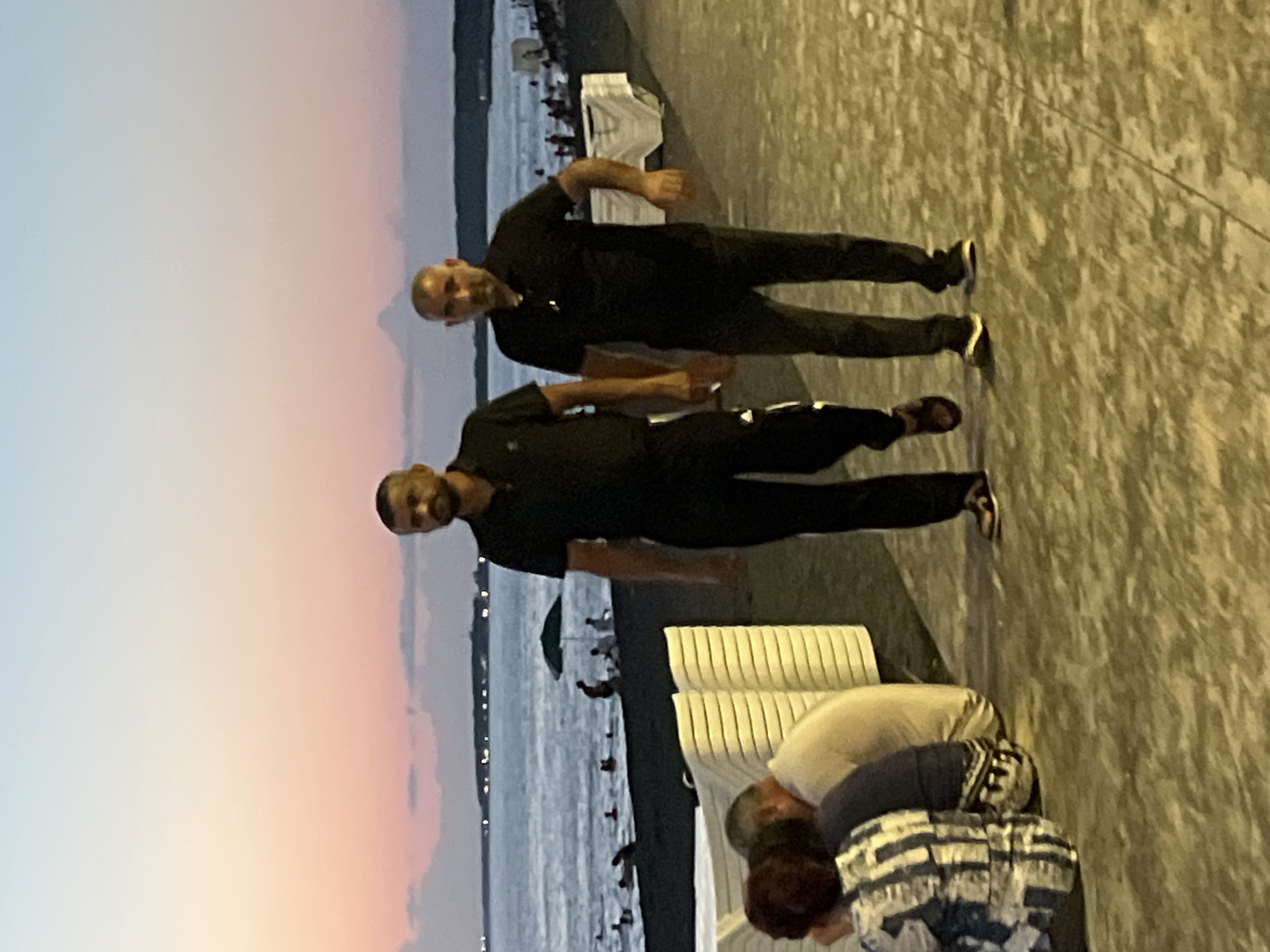 Two men in black shirts and pants are walking on a paved promenade along the Black Sea coast in Ureki, Georgia. The sky exhibits a gradient of light blue to pink, indicating dusk. Several beachgoers are visible in the distance, some in the water and others sitting on the dark sand, with a green umbrella providing shade. In the foreground, on the left, a group of people are seated, with one person wearing a black and white patterned shirt. Stacked white lounge chairs are also present along the promenade.
