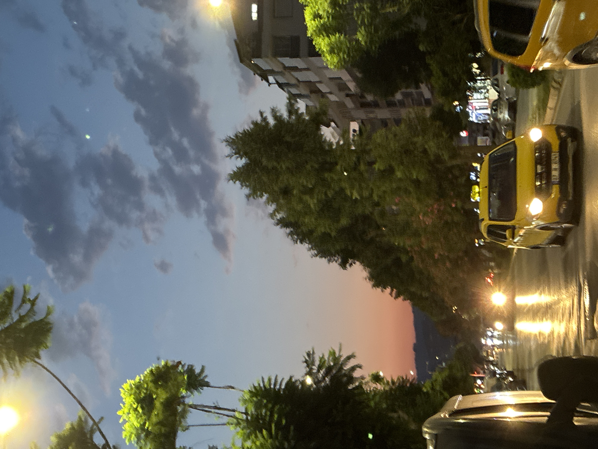 A street scene at dusk in Alanya, Turkey, shows a road lined with trees on both sides. A yellow taxi is prominent in the foreground, its headlights illuminated. Other vehicles are visible on the wet road, reflecting the lights of the city. The sky above is a mix of blue and orange hues, with clouds scattered across it. Buildings can be seen on the right side of the image, suggesting an urban environment. The overall impression is one of a busy street at the end of the day.