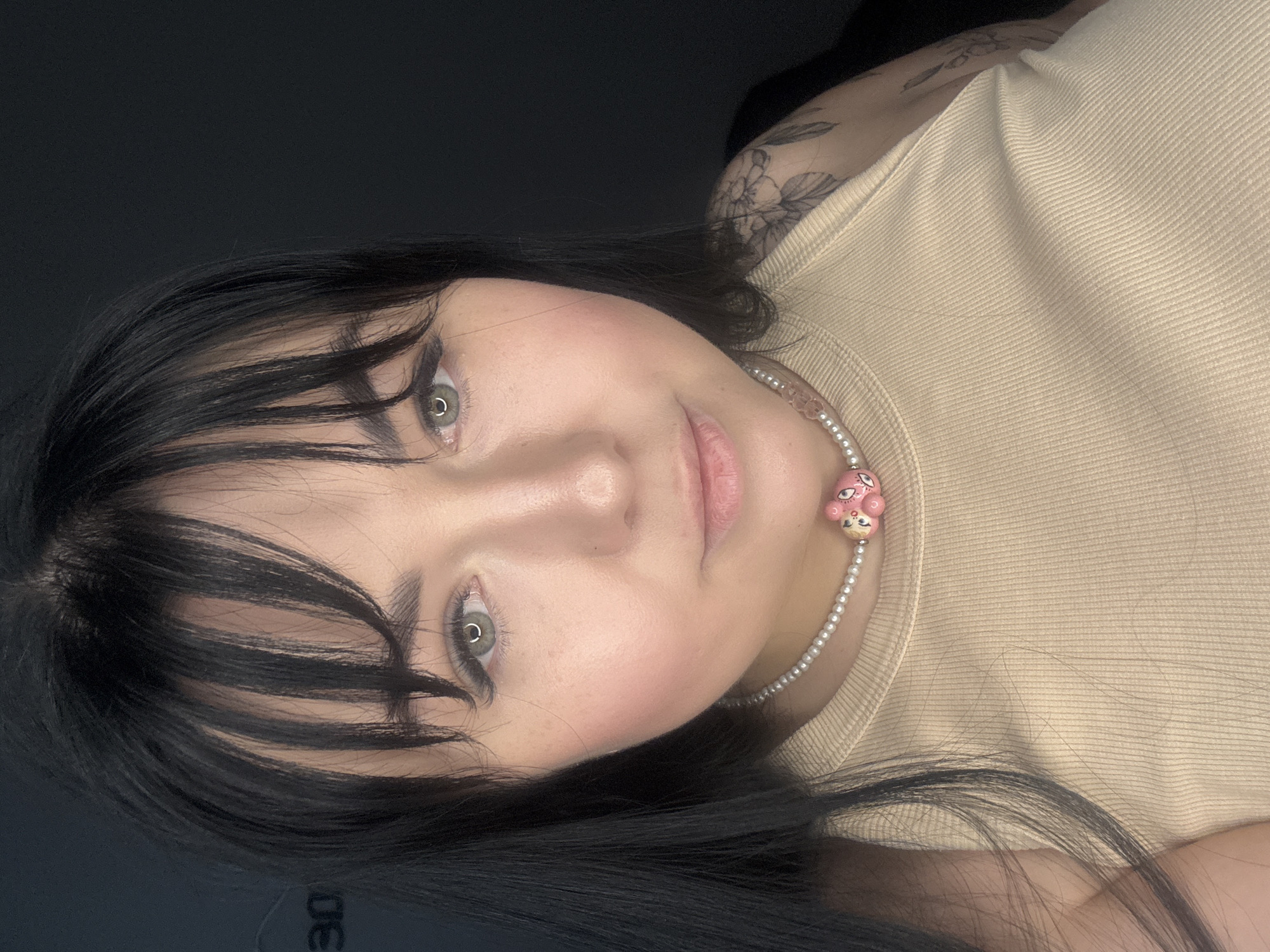 A close-up studio shot shows a woman with light skin, gray eyes, and black hair. She has short, choppy bangs and wears a beige ribbed top. A pearl necklace with a pink bear pendant adorns her neck. A tattoo of flowers is visible on her upper arm. The background is dark gray. The photograph was not taken in Chaikovsky, Russia.