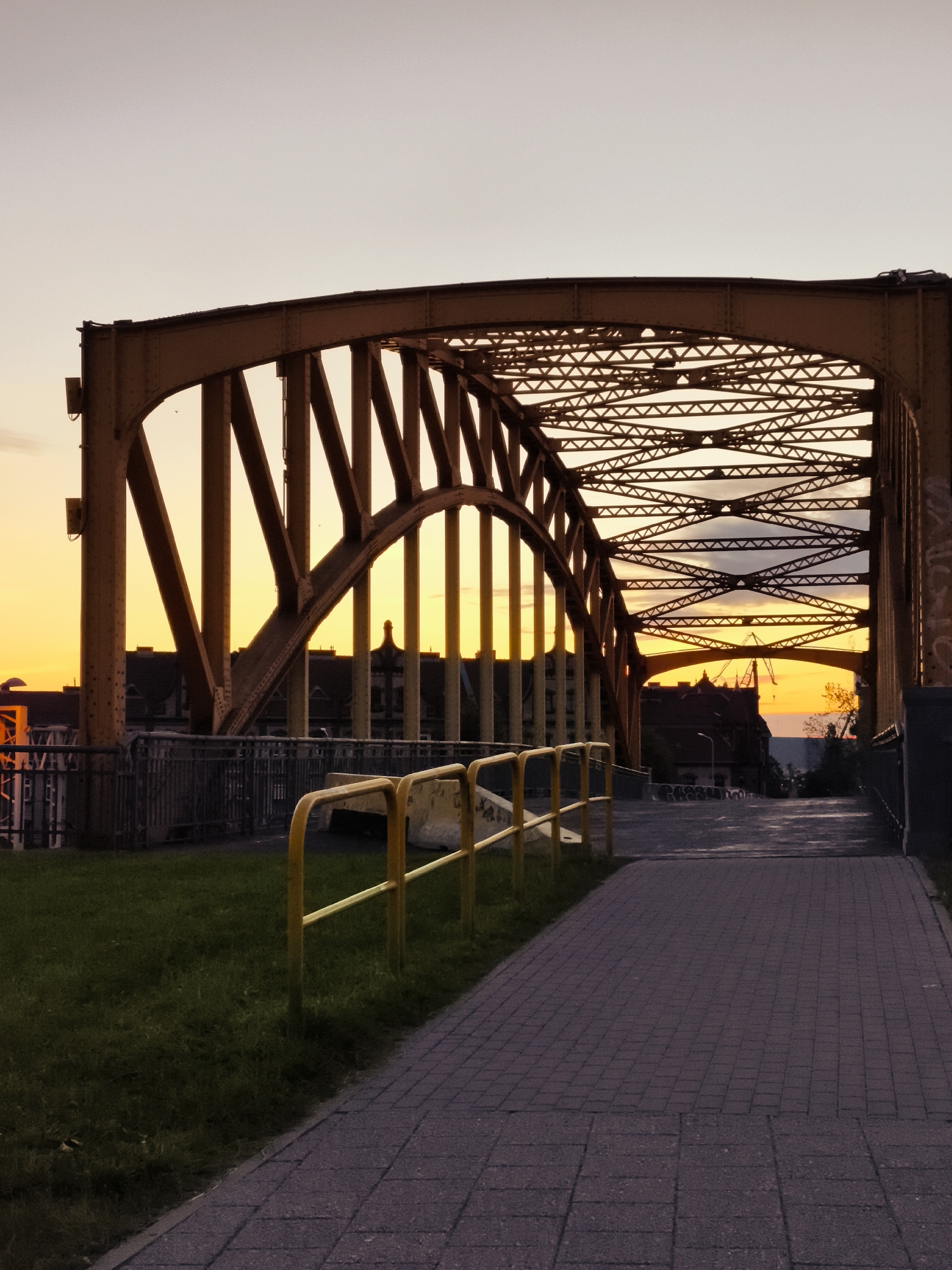 The image shows a pedestrian walkway next to a yellow metal truss bridge at sunset. The walkway is paved with grey brick pavers and bordered by a grassy area with a yellow metal railing. The bridge's intricate metal structure is visible against the twilight sky, which is transitioning from a pale orange to a soft grey. In the background, silhouetted buildings and a construction crane can be seen. The scene is devoid of people. This appears to be the Spichrzów Bridge (Most Stągiewny) in Gdańsk, Poland, a historic iron bridge that spans the Motława River.
