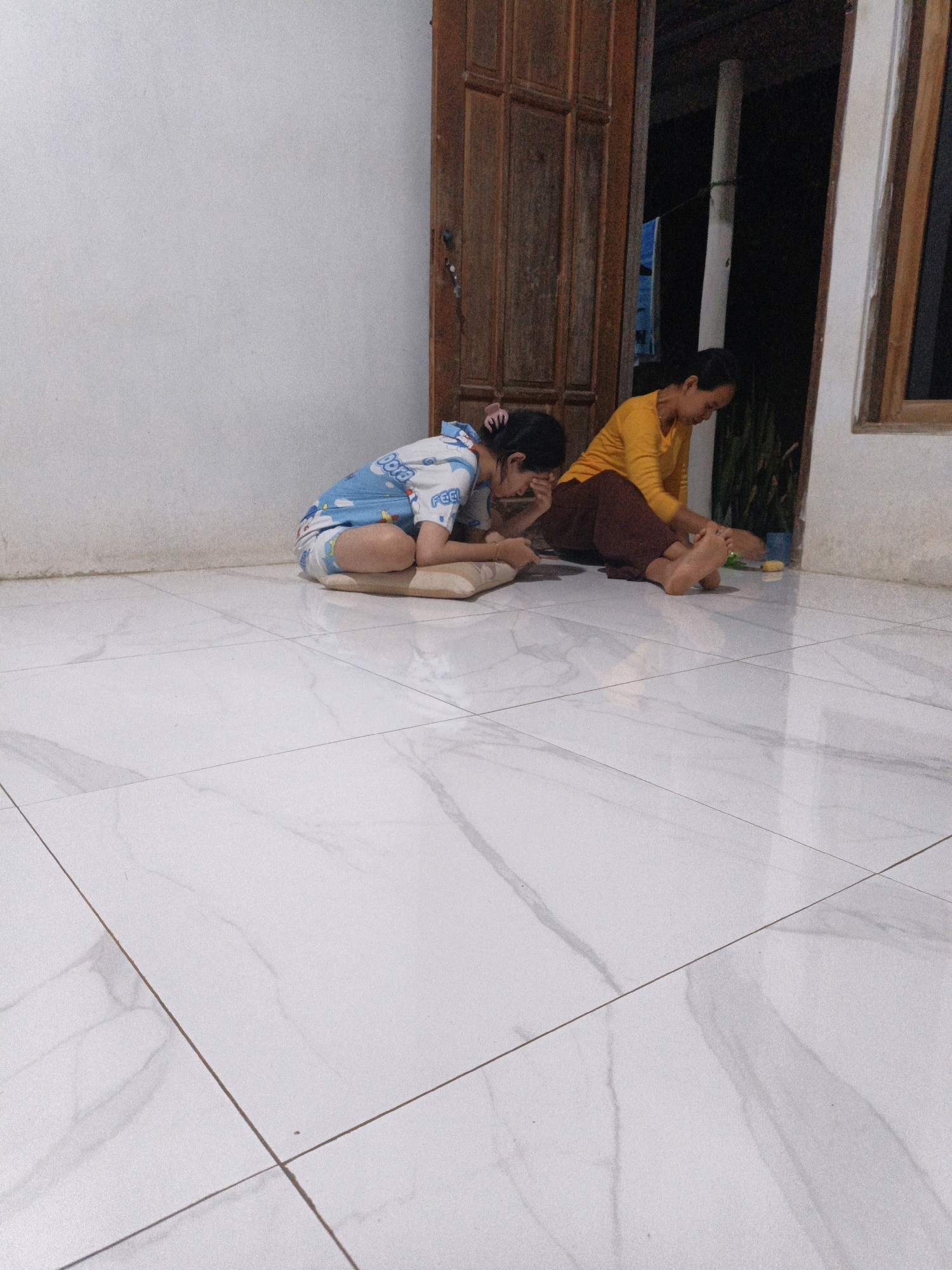 Two individuals are seated on a white tiled floor in what appears to be an indoor area with a wooden door and a window. The person on the left, a young girl, is wearing blue and white pajamas with a cartoon print and is sitting with her knees tucked towards her chest, resting her head in her hands. To her right, an adult woman is wearing a yellow long-sleeved shirt and brown pants, leaning forward with her hands on the floor. The environment suggests a domestic setting, possibly a home in Suwawal Timur, Indonesia, given the architectural style and climate. The lighting is dim, indicating it might be evening or nighttime.