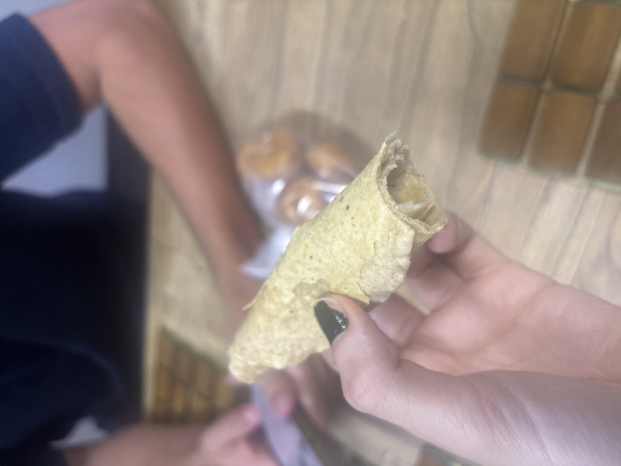 A person holds a partially eaten, rolled taco (flauta) in their right hand. The fingernails on the right hand are painted dark green. A second person, wearing a dark blue shirt, is partially visible to the left of the frame. In the background, there are cylindrical objects, possibly tamales, stacked in rows, and a plastic bag containing round objects, possibly donuts or rolls. The setting appears to be indoors, on a wooden surface. There is no information to deduce a specific location in Buenavista, Mexico.