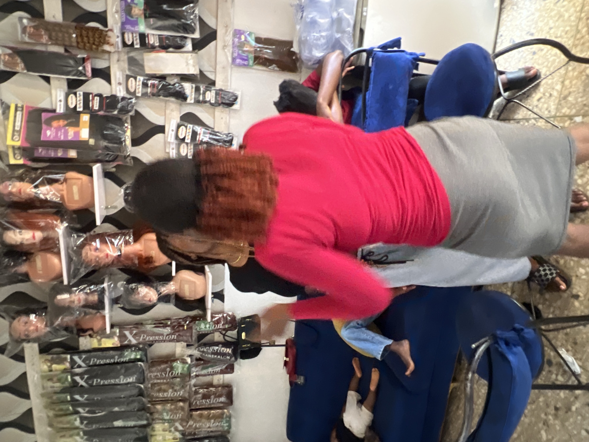 The image depicts the interior of a hair salon in Jos, Nigeria, filled with various hair products. Several racks of synthetic hair extensions, wigs, and mannequins with hairstyles are displayed on the walls. In the foreground, a person wearing a bright pink top and a grey skirt is interacting with another individual. To the left, a blue sofa is visible, with two individuals, one appearing to be a child wearing white shorts and the other a person in blue jeans, resting on it. The overall environment suggests a busy retail setting for hair extensions and related products.