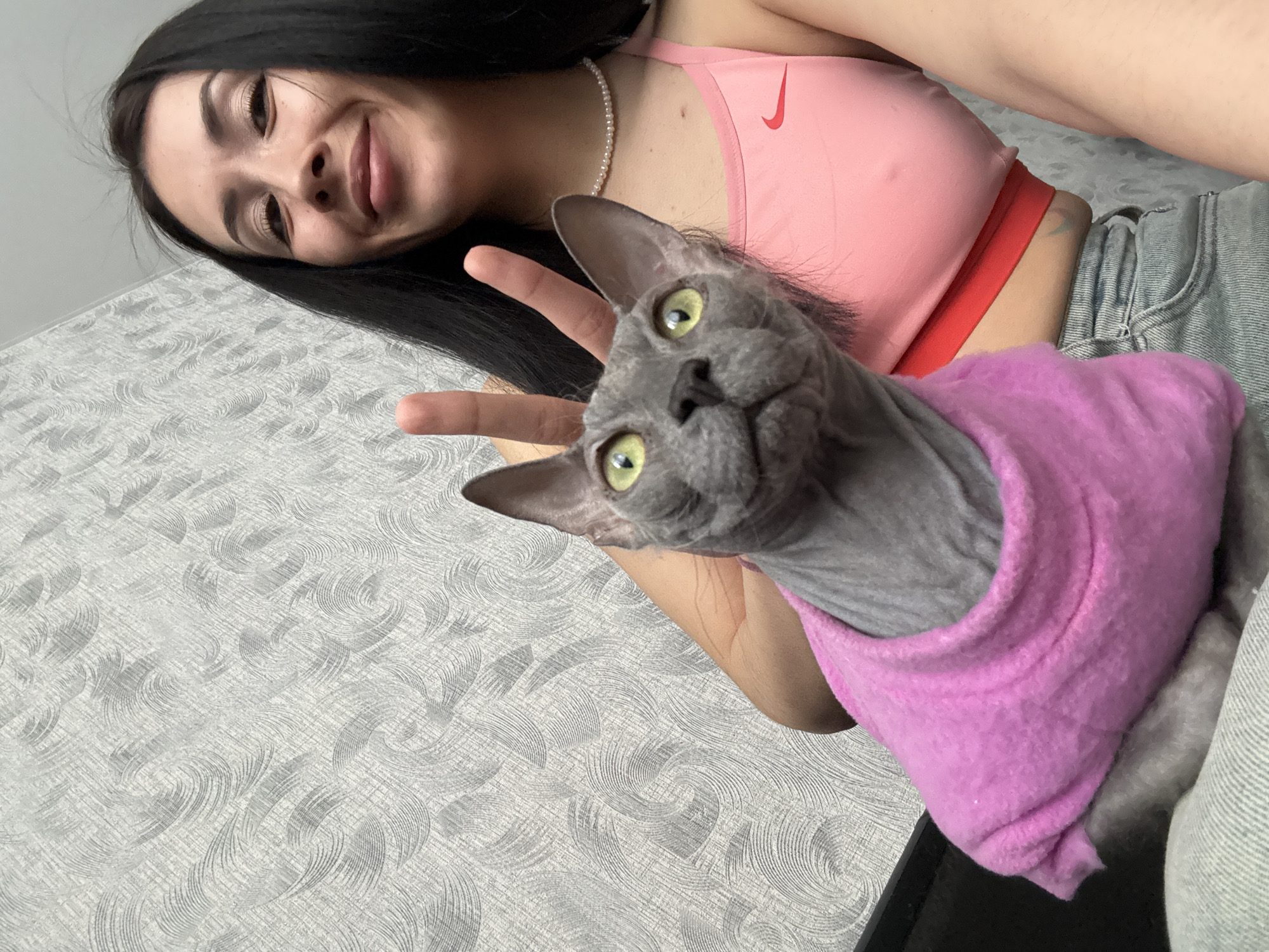 A young woman with long dark hair and a pearl necklace is posing with a gray Sphynx cat wearing a pink sweater. The woman is wearing a pink athletic top with a red band and light-wash jeans. She is smiling and holding up a peace sign with her left hand. The background is a gray patterned surface. The location is not discernible from the image.