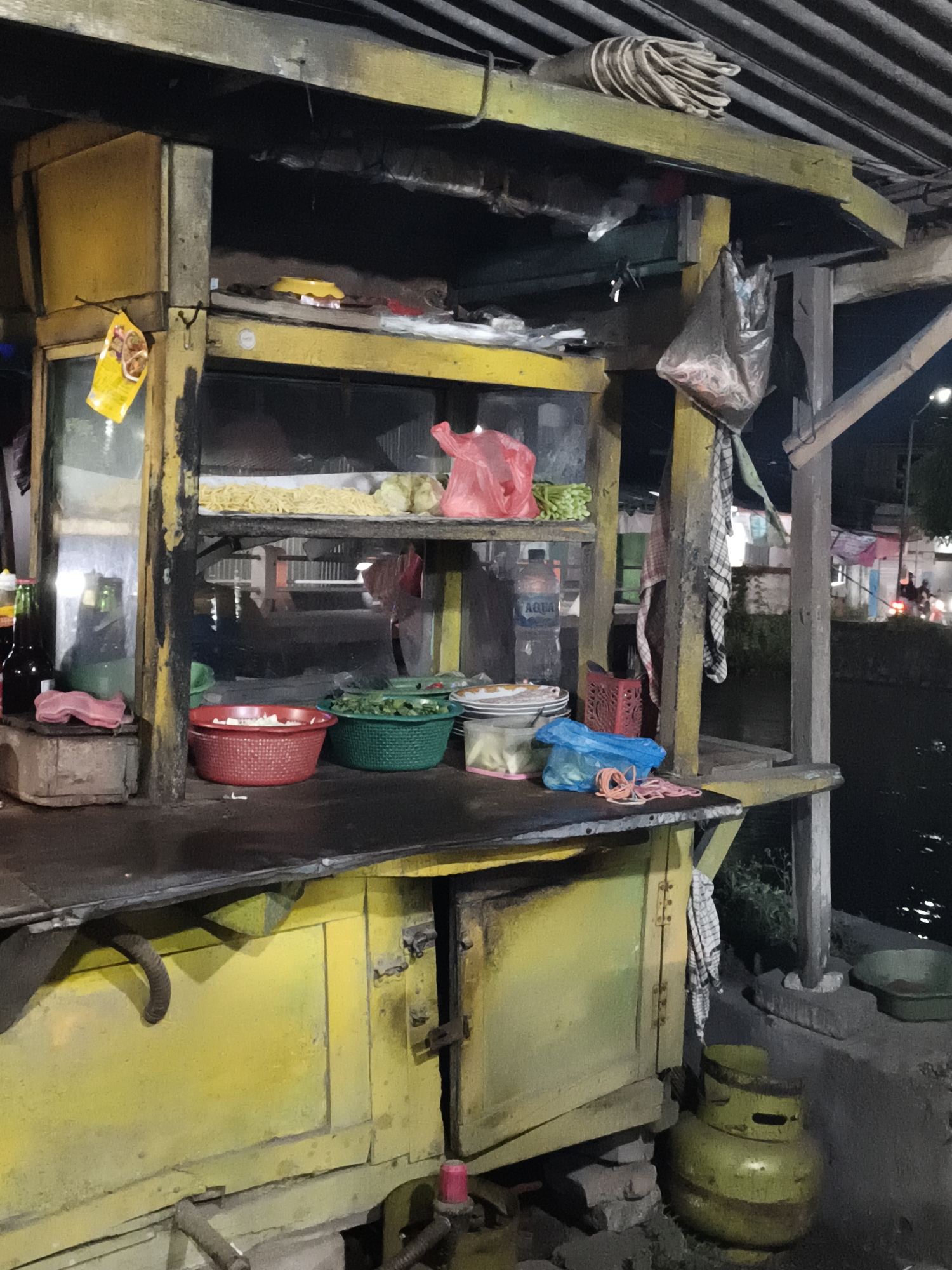 This is a nighttime, outdoor, medium shot of a food stall in Krikilan, Indonesia. The stall is constructed from weathered yellow wood and has multiple shelves displaying various food items and supplies. On the top shelf, there are bundles of what appear to be dried noodles or straws, and further back, some green vegetables. Below this, a plastic bag containing more green vegetables is visible.

The middle shelf holds two plastic baskets, one red and one green, both filled with chopped vegetables. To the right of these, a stack of white plates and a yellow plastic container are present. A bottle of "AQUA" mineral water is also on this shelf.

The counter area below displays more ingredients and kitchenware. A red plastic basket is on the left, with a pink cloth next to it. In front of the stall, a yellow propane tank sits on the ground, with some construction debris and a green bowl nearby. The overall scene suggests a humble, functional food stall operating after dark. The lighting is dim, with artificial lights from the stall and distant background lights illuminating the scene. The presence of the propane tank and the general state of the stall suggest it is a street food vendor.