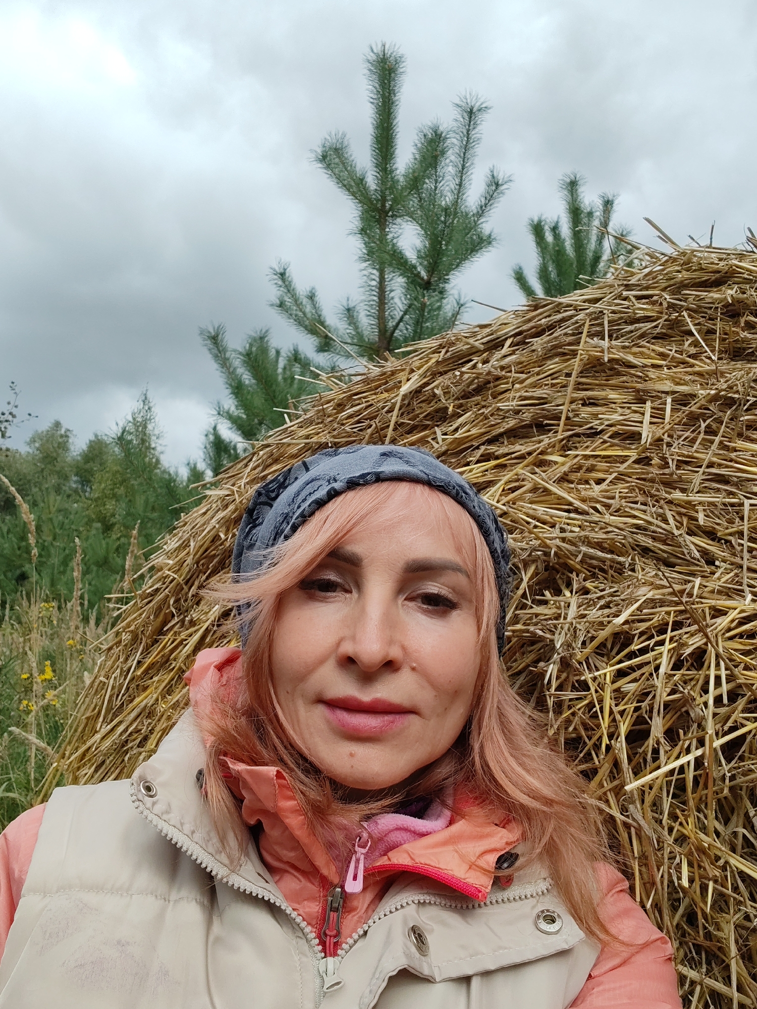A medium shot selfie features a middle-aged woman with pink hair standing in front of a large haystack and a pine tree. She is wearing a grey patterned headscarf, a light-colored vest over a pink jacket, and a pink undershirt. The woman is looking at the camera with a slight smile. The background consists of a dense haystack, a prominent pine tree, and green foliage. The sky is overcast and grey. The scene is likely set in an outdoor rural environment.