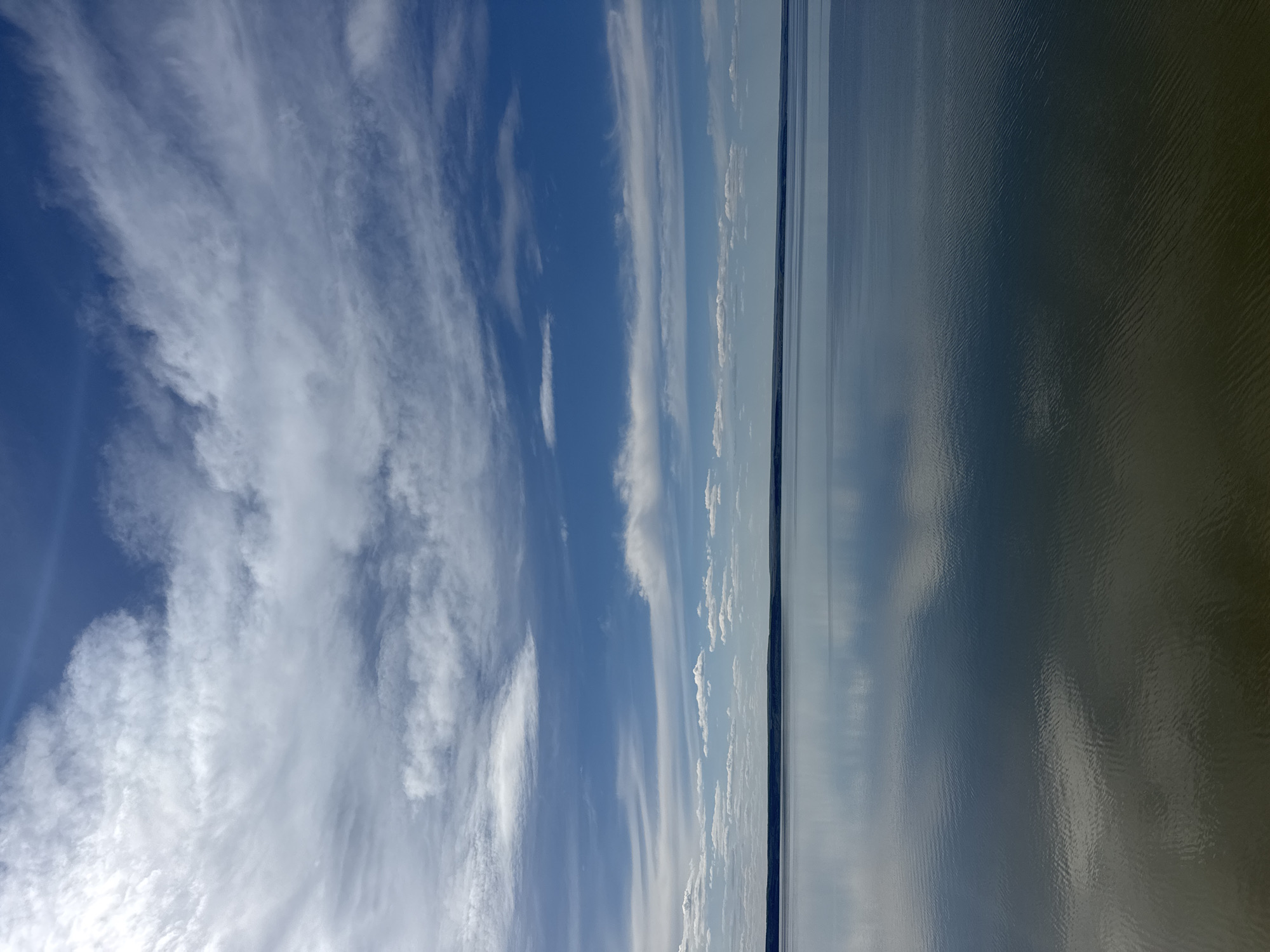 The image depicts a body of water under a blue sky with streaky, cirrus-like clouds. The water is calm with a slight ripple effect visible on the right side of the frame. A dark, thin landmass is visible on the horizon line, separating the sky and water. The scene appears to be a wide, open view of a lake or ocean on a clear day. The image is taken in Unknown, Unknown.