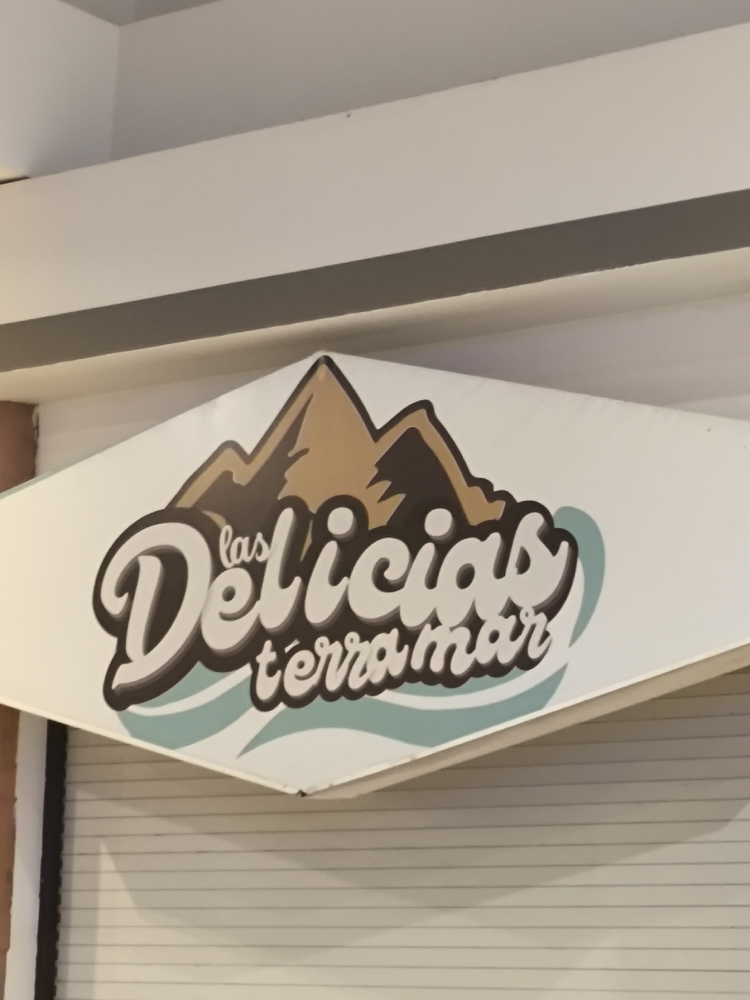 A sign for "las Delicias terramar" is mounted on a white diamond-shaped surface, with the bottom section a silver-colored roll-up garage door. The sign features a stylized drawing of mountains in shades of brown. The location is likely in the city of Puebla, Mexico based on the name of the business.