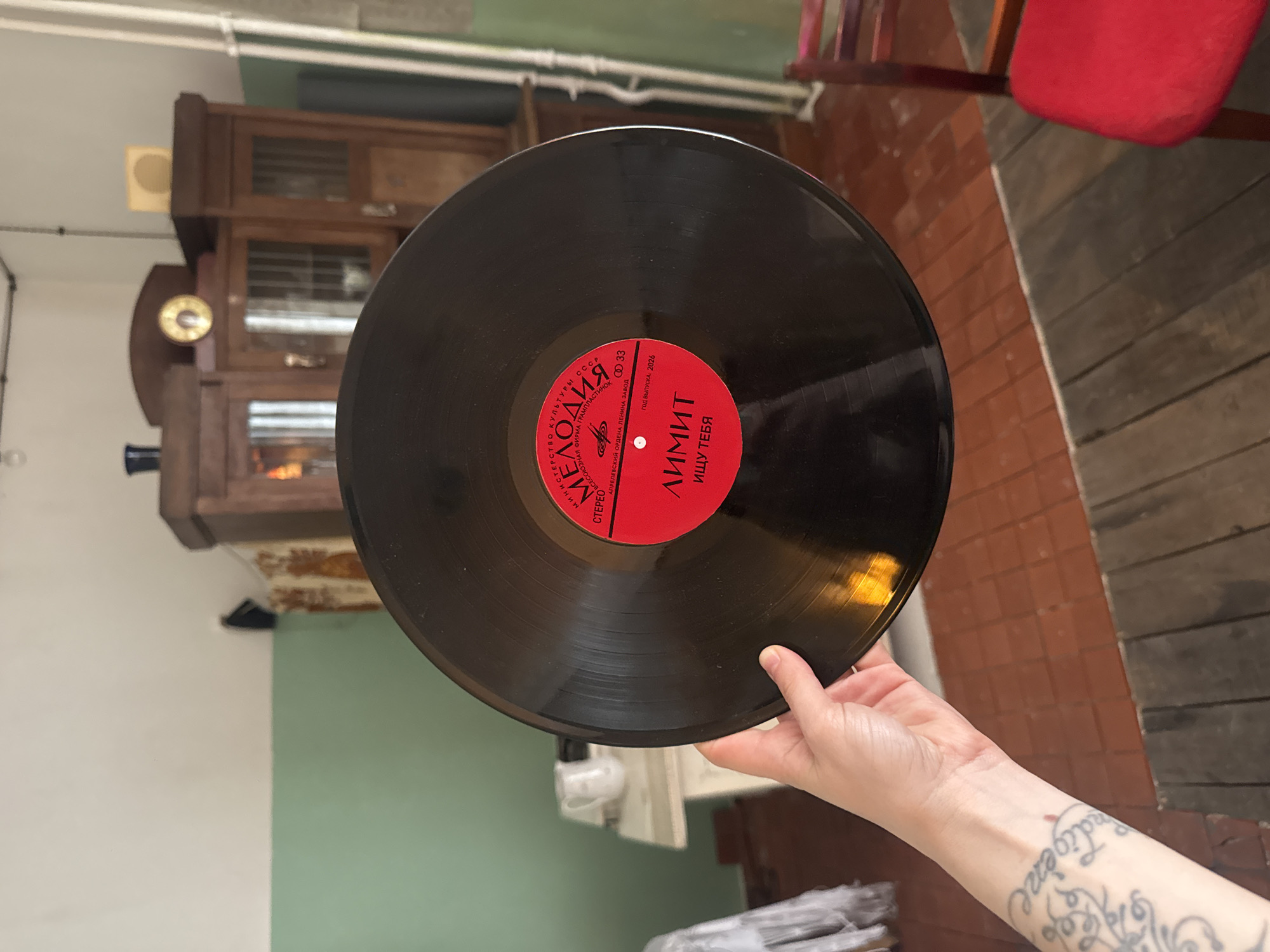 A person holds a black vinyl record with a red label in front of a brick and wood-floored room. The record has "MELODIA" printed in Cyrillic on the left side of the label, and "ЛИМИТ" (LIMIT) and "ИЩУ ТЕБЯ" (I'm Looking for You) printed on the right. The wrist of the person holding the record has a tattoo in black ink. The background includes an antique wooden cabinet with glass doors, a red chair, and a wall painted white and green. The location is likely Moscow, Russia, given that "MELODIA" was a Soviet record label.