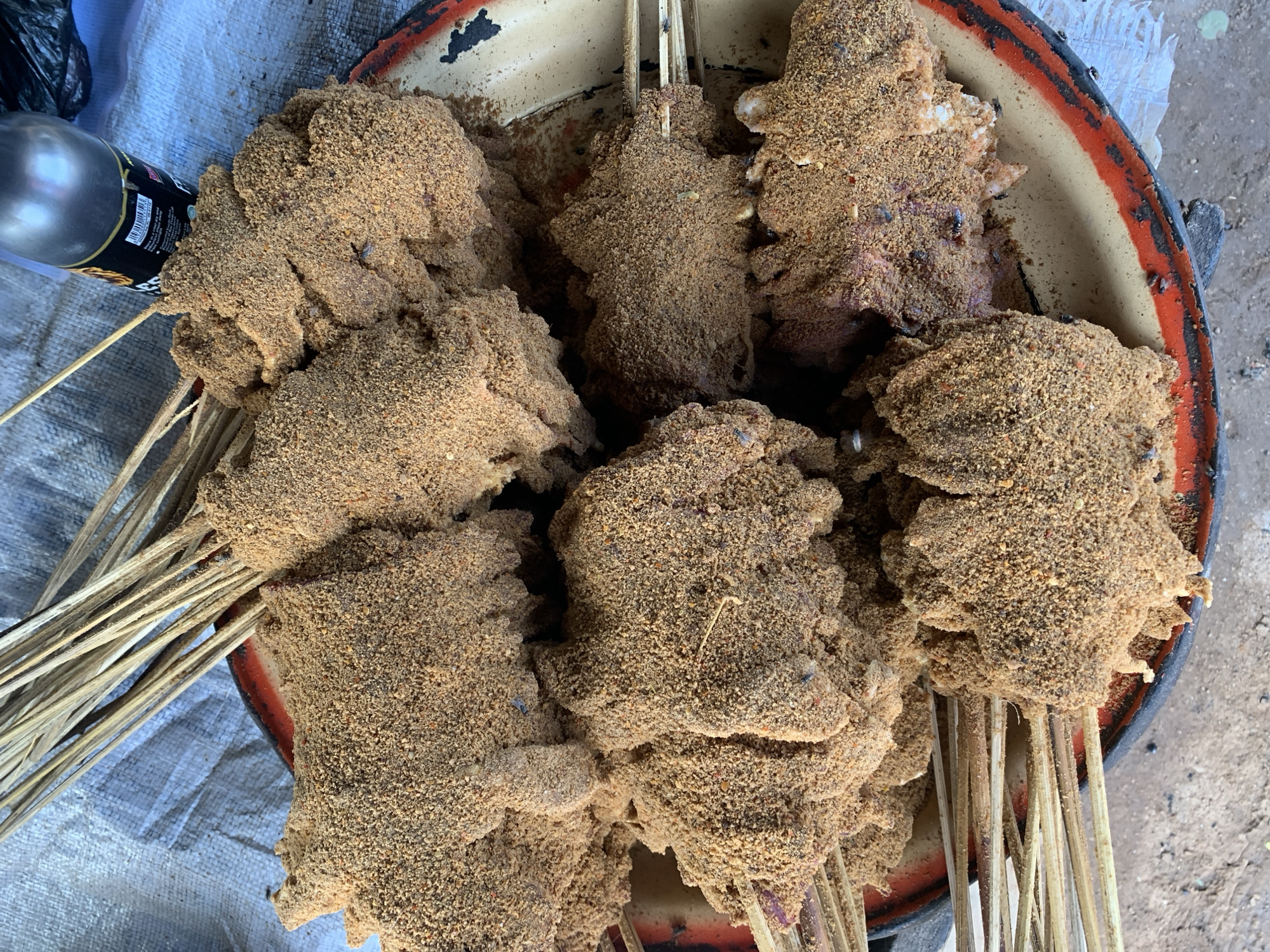 The image shows a close-up view of a metallic bowl filled with several skewers of meat, heavily coated in a brown, grainy spice rub. The skewers are resting within a weathered, round bowl with a faded red and black rim. To the left, a partial view of a silver bottle with a black label is visible. The backdrop includes a weathered surface, perhaps a table or ground covering. The scene is identified as New Stadium, Nigeria.