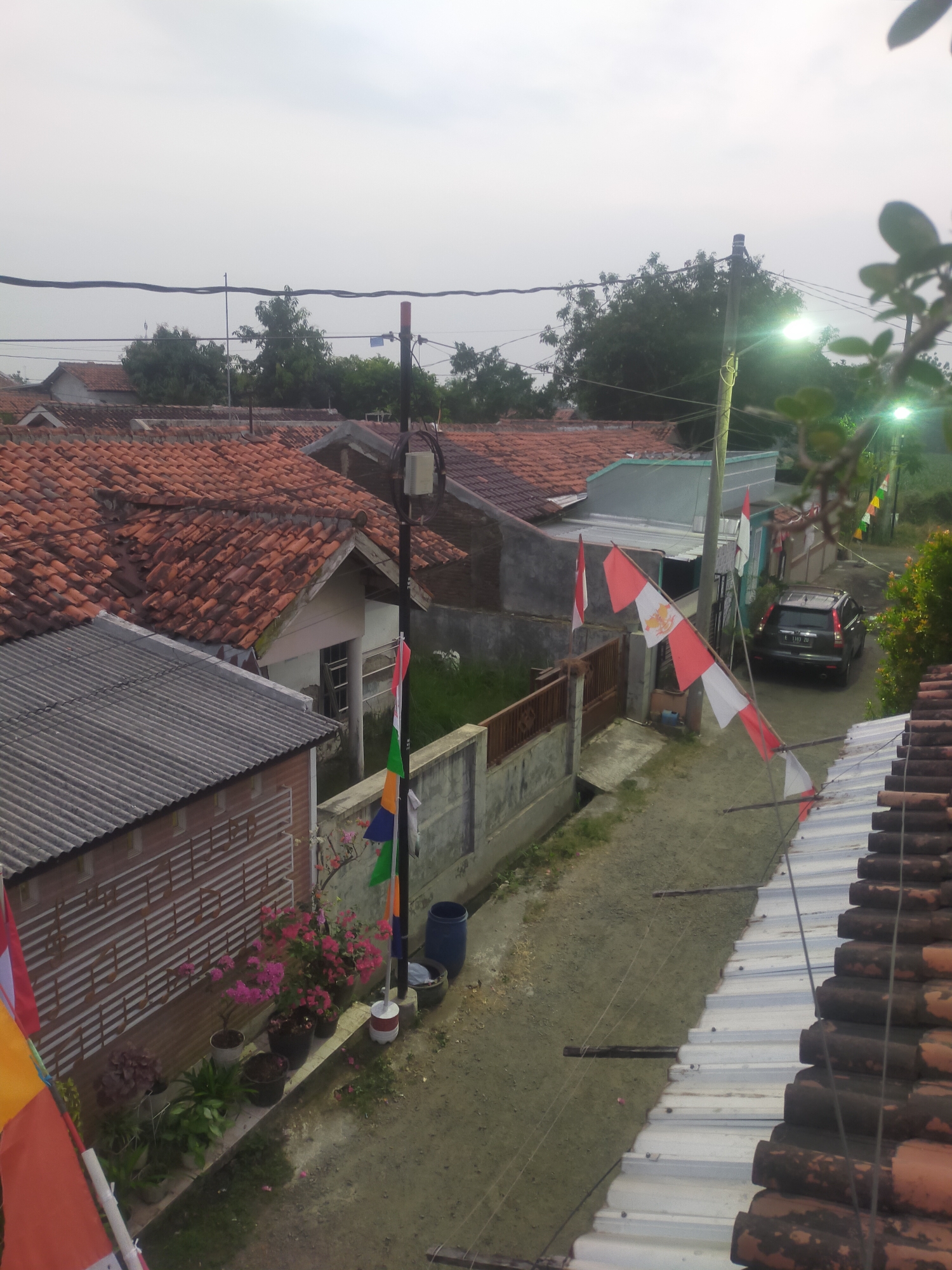 The image is a high-angle shot of a residential street in Tegalandong, Indonesia, possibly during daytime with overcast skies. The foreground features the edge of a tiled roof and a corrugated metal roof. In the midground, a narrow dirt road winds through the scene, flanked by houses with red tile roofs and concrete walls. Several Indonesian flags, red and white, are displayed on poles along the street. A dark-colored SUV is parked on the side of the road. Potted plants, including flowering bushes, are visible along a low wall. Overhead power lines crisscross the sky. The overall environment appears to be a somewhat modest, possibly rural or suburban, Indonesian neighborhood.