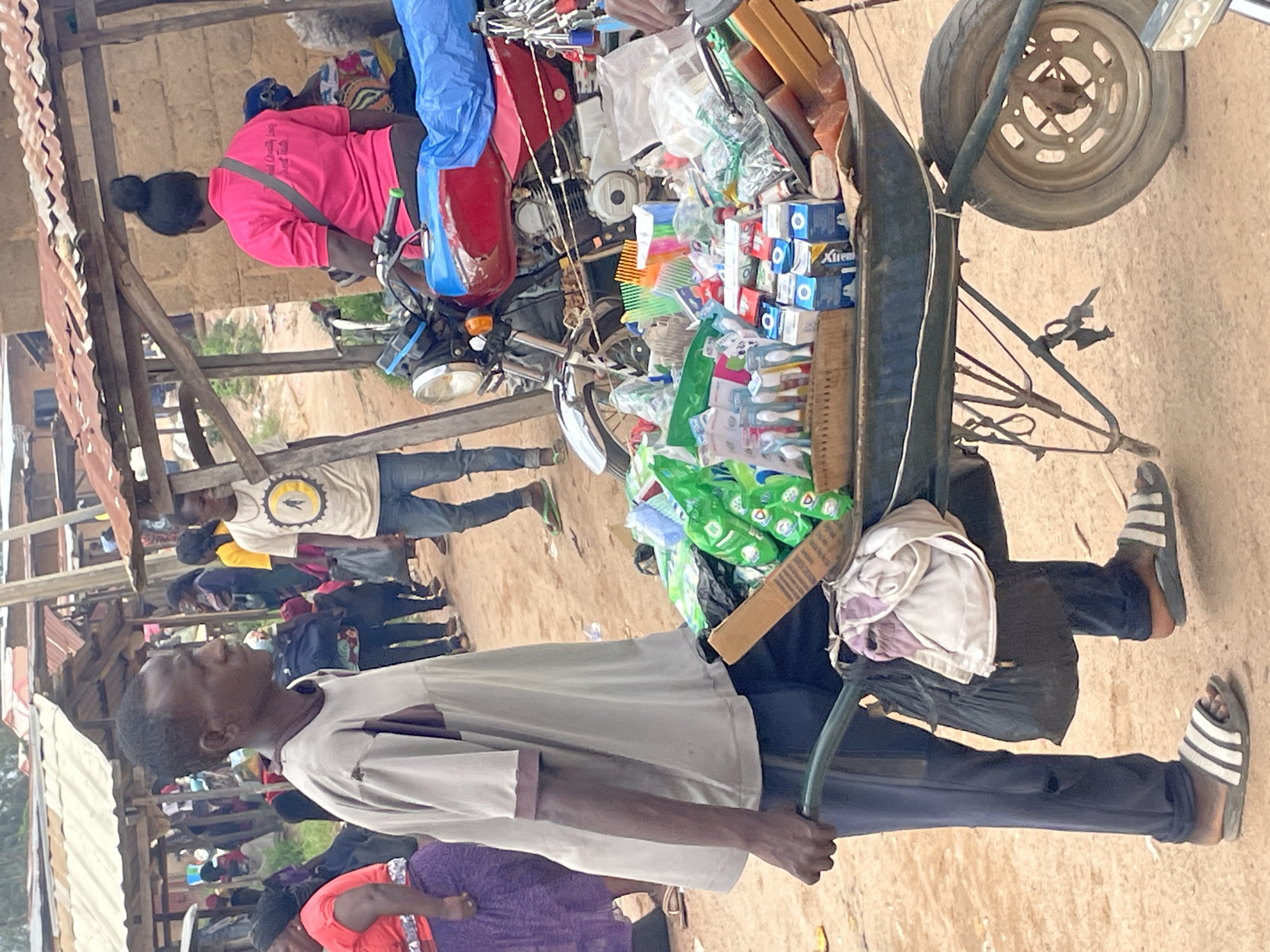 In Wang, Nigeria, a man walks with a wheelbarrow filled with assorted goods, including hygiene products like toothbrushes and soap, as well as various packaged items. He is wearing a grey t-shirt with brown trim and dark blue pants, along with striped sandals. The wheelbarrow is laden with merchandise, some of which is in green packaging, and a larger black bag hangs from the side. In the background, a motorcycle is parked, and other people are visible in a market setting with makeshift stalls constructed from wood and corrugated metal. A woman in a pink t-shirt with a ponytail is seated on the motorcycle. The ground is dusty earth.