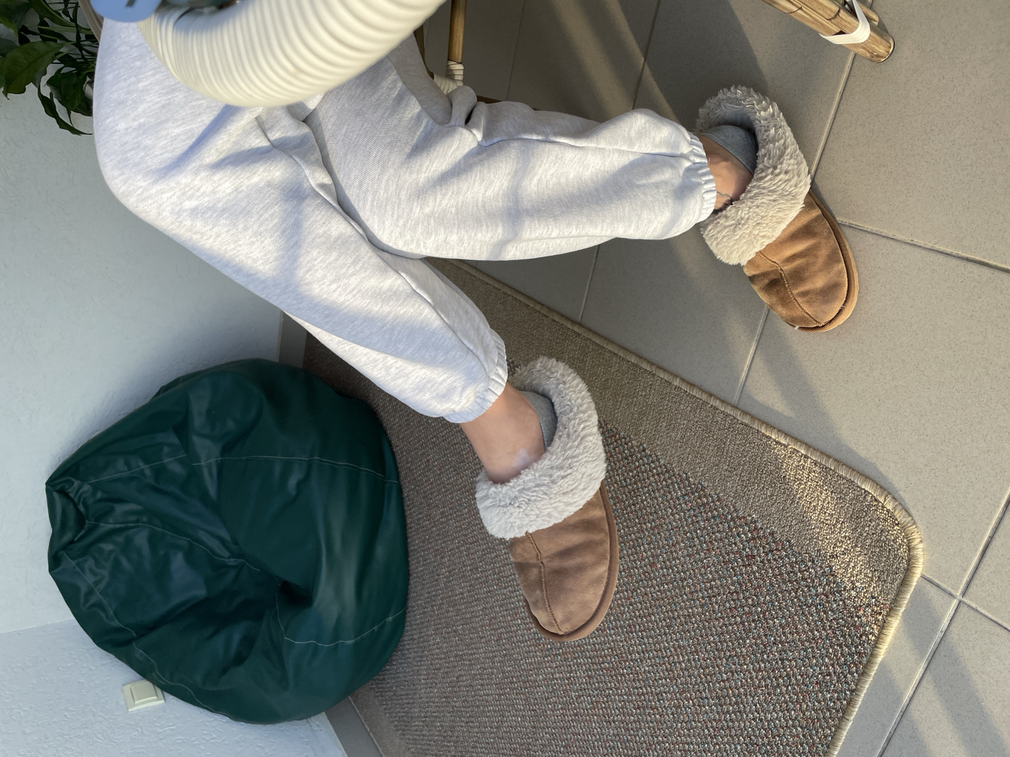 A person wearing light gray sweatpants and brown suede slippers with a fur lining is sitting in a room. A green beanbag chair is visible next to the person's left foot. The floor is tiled and partially covered with a neutral-toned rug. The location is potentially Rafailovići, Montenegro. A white, coiled tube is partially visible above the person. A wooden chair leg is visible in the background.
