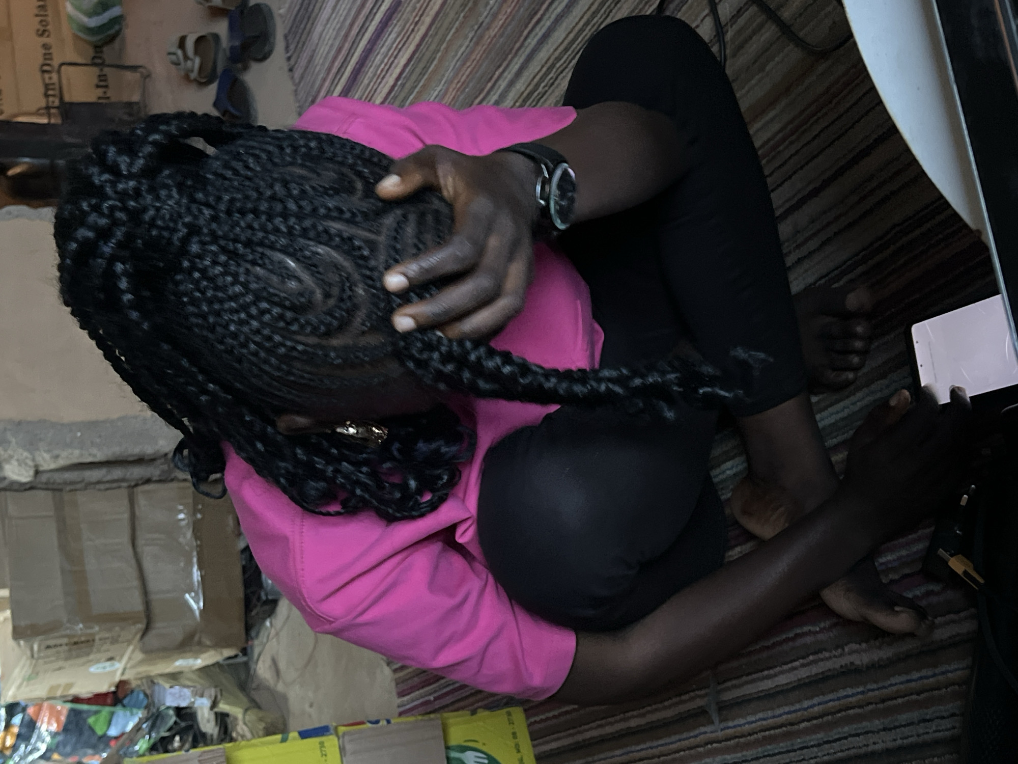 The image depicts a person sitting on a striped carpeted floor, with their head bowed and hand covering their face. They are wearing a bright pink t-shirt and black pants, with a watch on their left wrist. The background shows stacked cardboard boxes, some with labels, and a wall with posters or printed material. Several pairs of sandals are visible in the background. A smartphone and a charging cable with a yellow connector are on the floor in front of the person. The scene suggests a storage area or a room with household items. The location is identified as Jos, Nigeria.